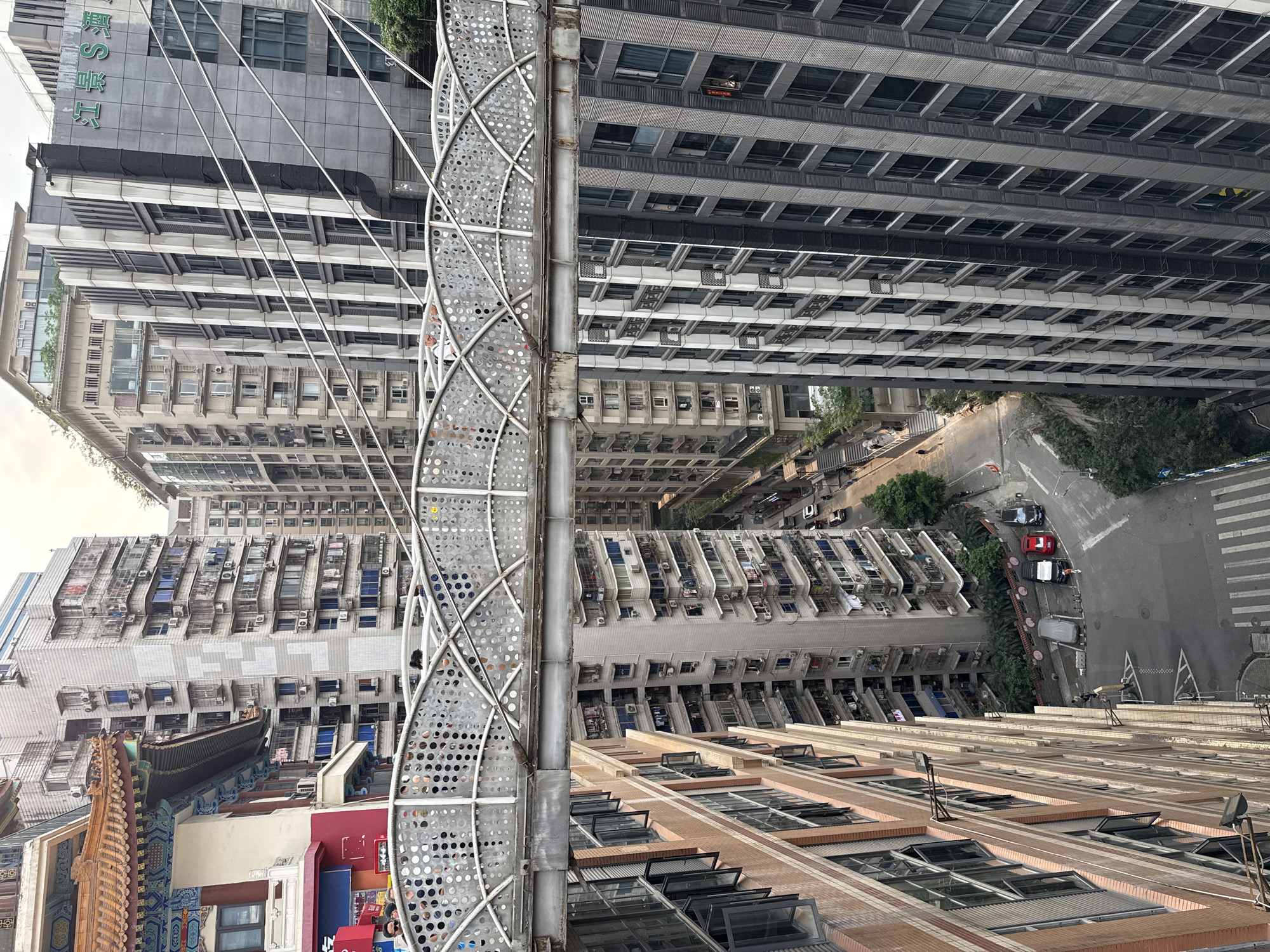 A high-angle, eye-level shot depicts a dense urban environment in Yuzhong District, China, characterized by a mix of architectural styles.
Several high-rise buildings dominate the skyline, constructed in a variety of concrete and brick facades. Many of these structures have rows of windows with protruding air conditioning units. An ornate, traditional Chinese building with a decorative tile roof and colorful details contrasts with the more modern constructions.
A light grey, metallic structure runs vertically through the center of the image. This appears to be a public art installation or architectural element with numerous small circular cutouts throughout its design.
A narrow street is visible between the buildings, showing cars parked along the side and a pedestrian crossing. The weather appears to be clear.