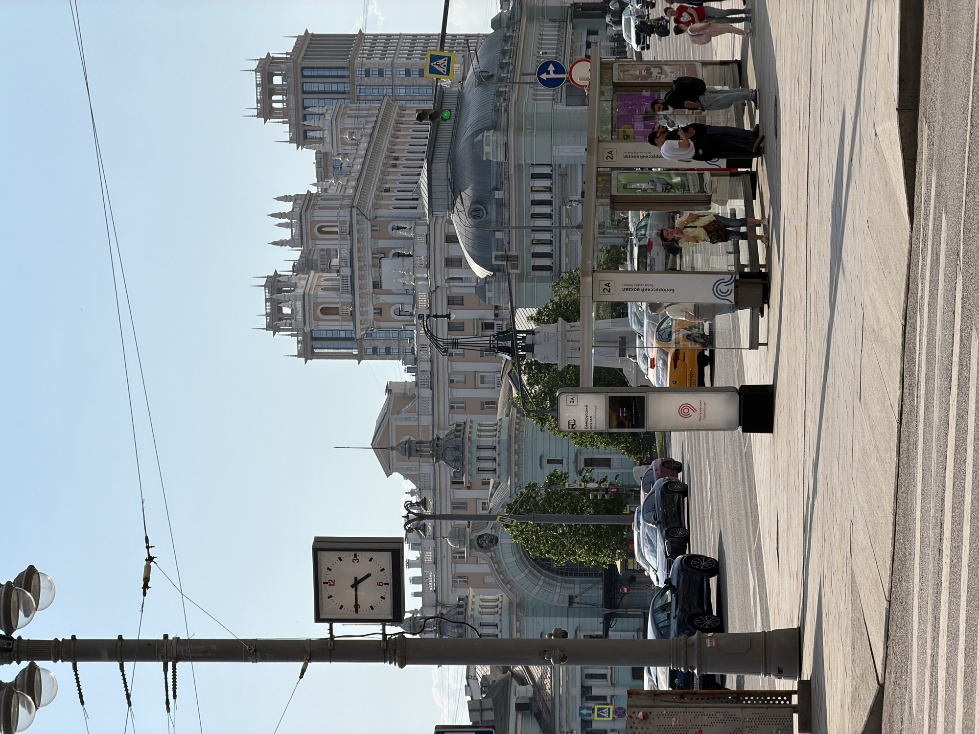 Here is a description of the image:
The image captures a street scene in Moscow, Russia. The architectural style of the buildings lining the street suggests a historic district. Notably, the buildings display ornate facades. A clock mounted on a pole indicates the time. Several cars are parked along the street, which appears to be an avenue. Pedestrians are visible on the sidewalk, suggesting the area is a public space. Additionally, street signs and advertisements are positioned along the walkway.