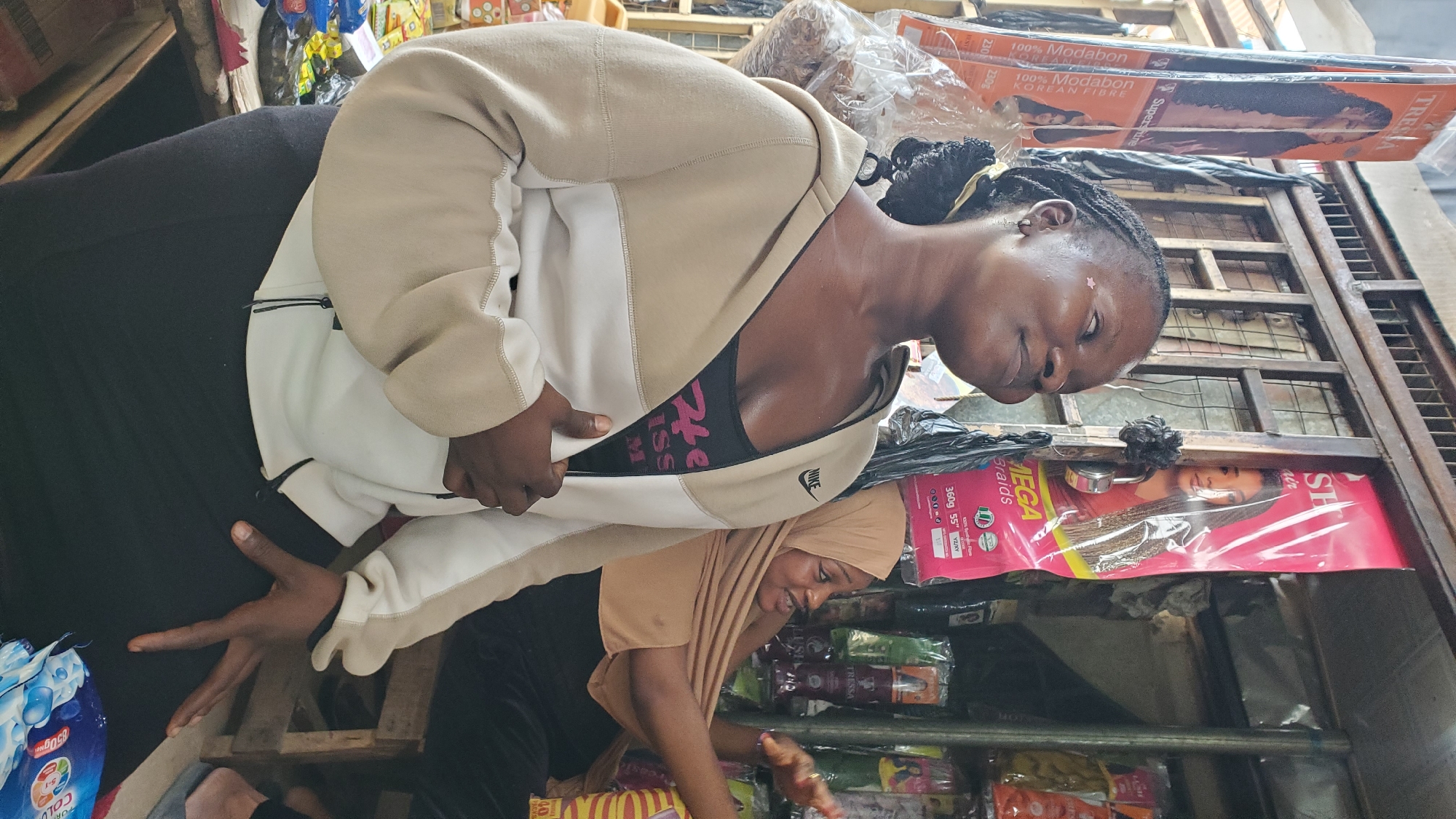 The image shows two women in an outdoor market stall in Jalingo, Nigeria. The woman in the foreground, facing left, is wearing a beige and white zip-up hoodie over a black tank top. She has dark braided hair pulled back and is looking upwards and to her left with a slight smile. There is a small pink star-shaped sticker on her cheek. The woman in the background, facing right, is wearing a tan hijab and a black outfit. She is gesturing with her right hand and appears to be speaking. The stall is filled with various hair products, including brightly colored packages of braids and weaves, displayed on shelves and hanging from the ceiling. The overall environment suggests a busy marketplace setting.