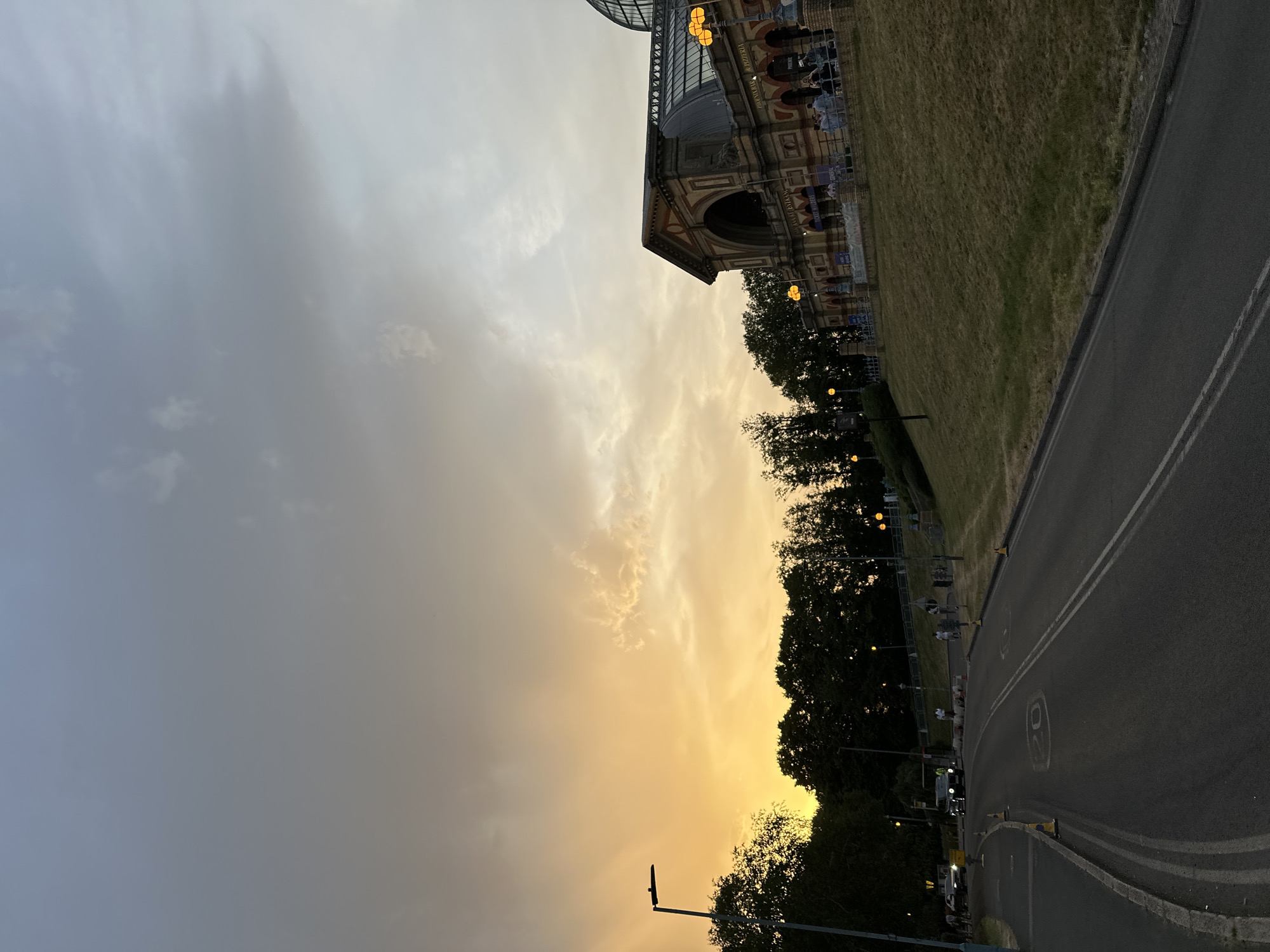 The image captures a street scene at twilight. On the right, a road with white lines is visible, leading towards a stretch of trees and streetlights. Beyond the trees, a building with a dome-like structure stands, partly illuminated. The left side of the frame is dominated by a cloudy sky, transitioning from dark gray to a golden hue near the horizon.