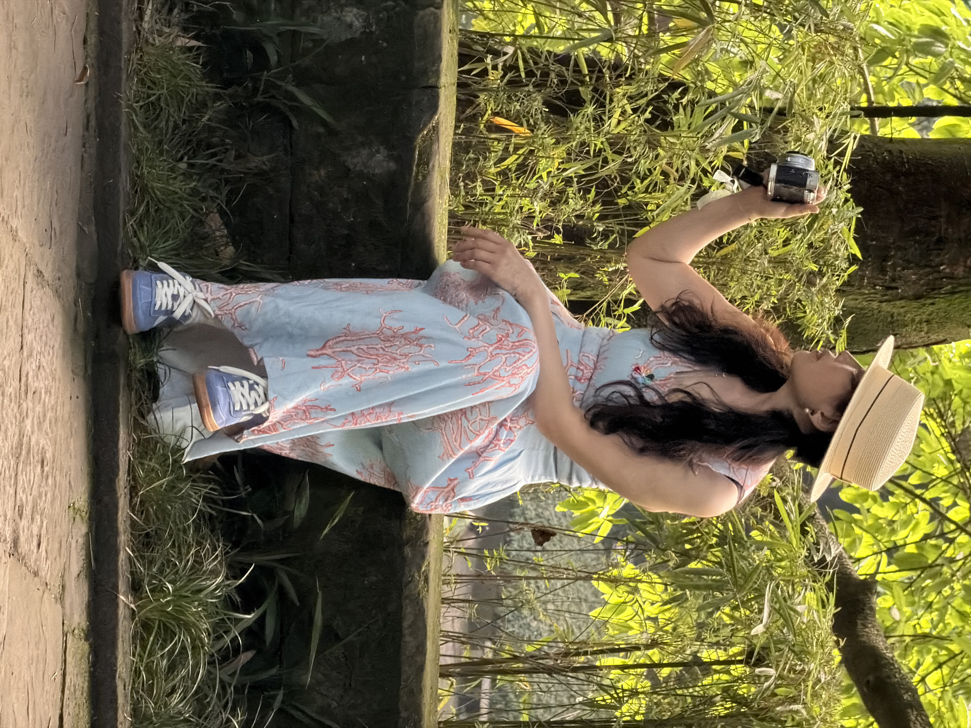 In Yuzhong District, China, a woman wearing a straw hat and a light blue dress with a coral pattern is seated on a stone ledge. She holds a vintage camera in her right hand, appearing to take a photo. The background consists of dense green foliage and a large tree trunk. The woman is wearing blue sneakers with white laces and an orange sole. The lighting suggests it is daytime.