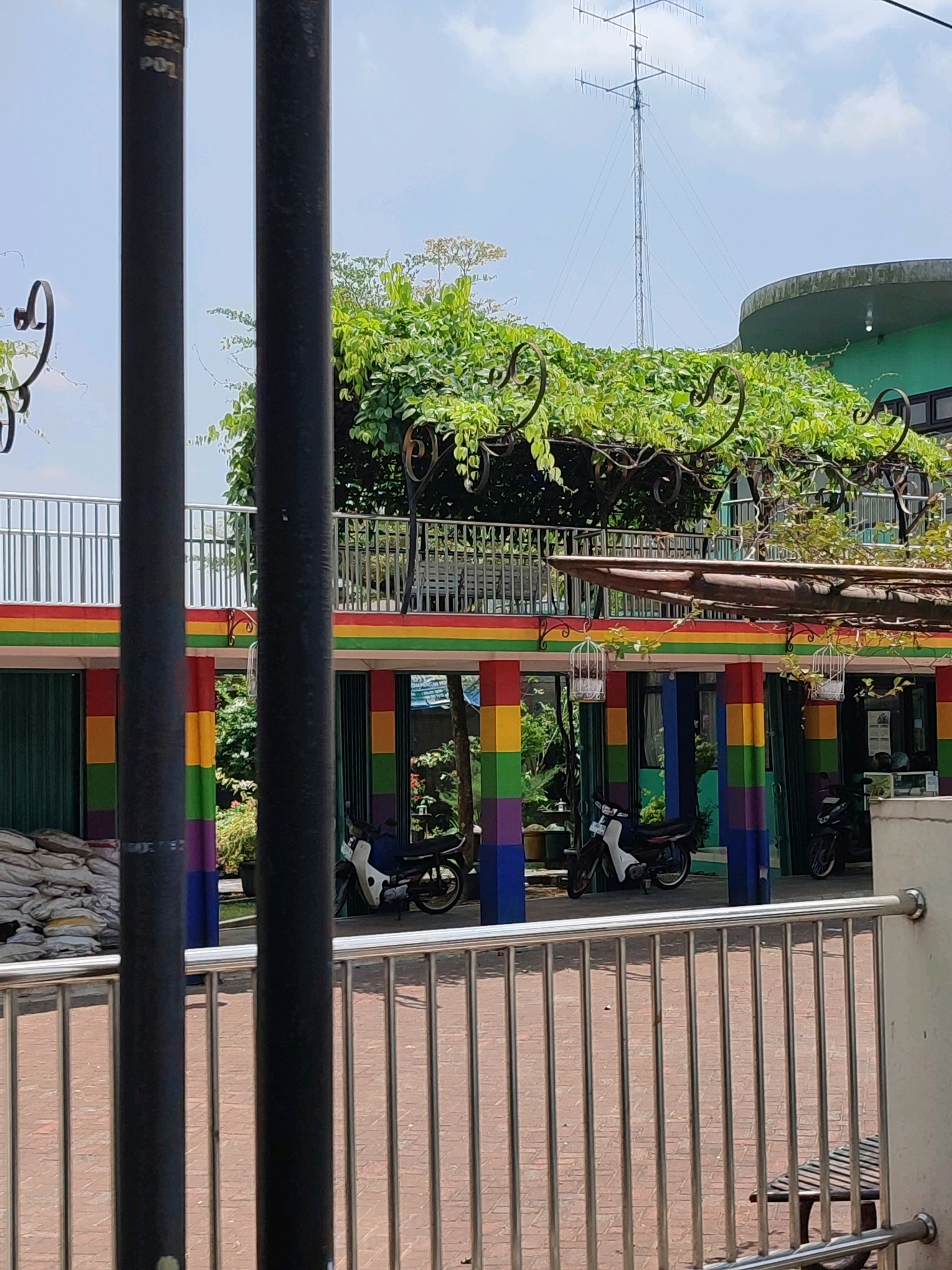 A medium shot captures an outdoor scene in Magelang, Indonesia, on a clear day. The foreground is dominated by two thick, black metal poles obscuring a view of a silver metal railing and a red brick-paved courtyard with a metal bench. Behind the railing, a building with a rainbow-colored facade stands out, featuring columns painted in red, yellow, green, and purple. Two motorcycles are parked in front of the building. Above the rainbow structure, a balcony with metal railings is covered by lush green vines and foliage. In the background, a tall radio tower rises against the sky, and the top of a rounded green building is visible. The scene appears to be public, possibly a shop or commercial establishment, given the facade and the presence of vehicles and seating. The overall impression is one of a vibrant and somewhat eclectic urban environment.