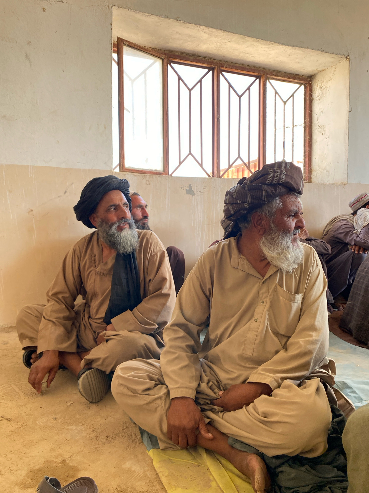An eye-level shot shows a group of men sitting on the floor of a room. The room has light-colored walls and a high ceiling. A large window with wooden panes and metal bars is visible in the background, letting in natural light. The men are dressed in traditional clothing, with turbans and loose-fitting garments. Two men are in the foreground, seated cross-legged and facing to the right. The man on the left has a black turban and a black scarf draped around his neck. He has a grey beard and is looking upwards. The man on the right has a brown and white striped turban and a long white beard, and he is looking to the side with a slight smile. Other men are visible further back in the room, also seated on the floor. The floor appears to be made of dirt or sand, with some fabric spread out on it. The overall impression is one of a gathering or meeting taking place in a modest setting. The location is identified as the city of Pasab, Afghanistan.