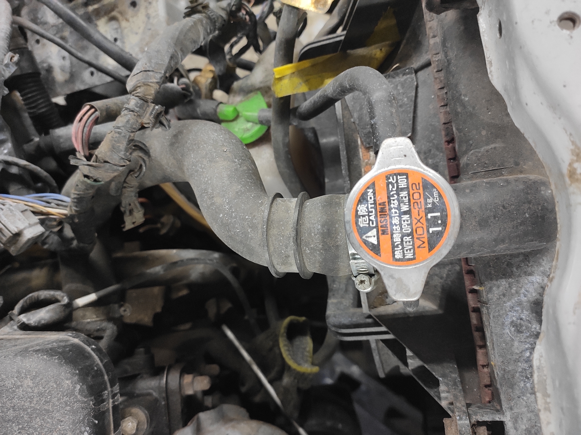 This is a close-up, eye-level, indoor photograph of a car engine bay, likely in a garage or workshop in Homyel, Belarus. The focus is on the radiator cap and the surrounding hoses and wiring. The radiator cap is silver with an orange and black label that reads "CAUTION" and "NEVER OPEN WHEN HOT," along with Japanese text and "MOX-202" and "1.1 kg/cm2". A grey rubber hose is clamped to the cap. Various other hoses, wires, and mechanical components are visible in the background, covered in dust and grime, suggesting the car may be older or in need of maintenance. The lighting is dim, and the image has a slightly grainy texture. No people are present in the scene.