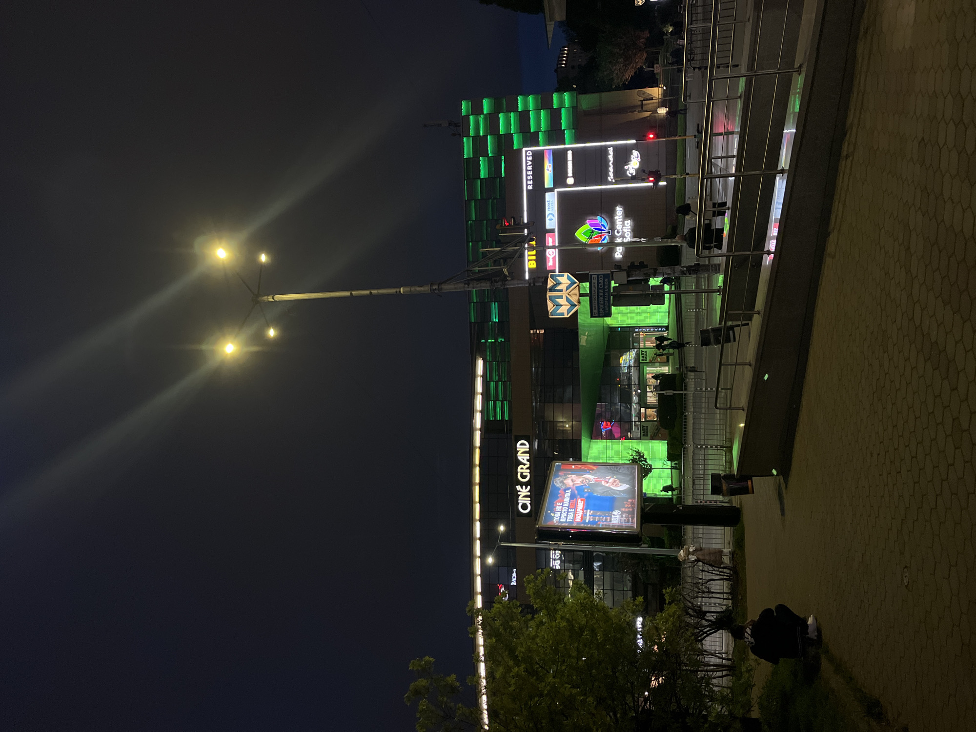 The image captures an evening scene in Sofia, Bulgaria. The BTC City Park Center Sofia is visible in the background, featuring a modern architectural style with green lighting accents. Adjacent to it, the "CINE GRAND" cinema is prominently displayed. A large billboard advertises "BELL" drink. Street lights illuminate the area, casting light on the paved walkway where pedestrians are present. The overall atmosphere suggests a contemporary urban environment. The Sofia metro logo is visible, indicating the presence of a nearby metro station.