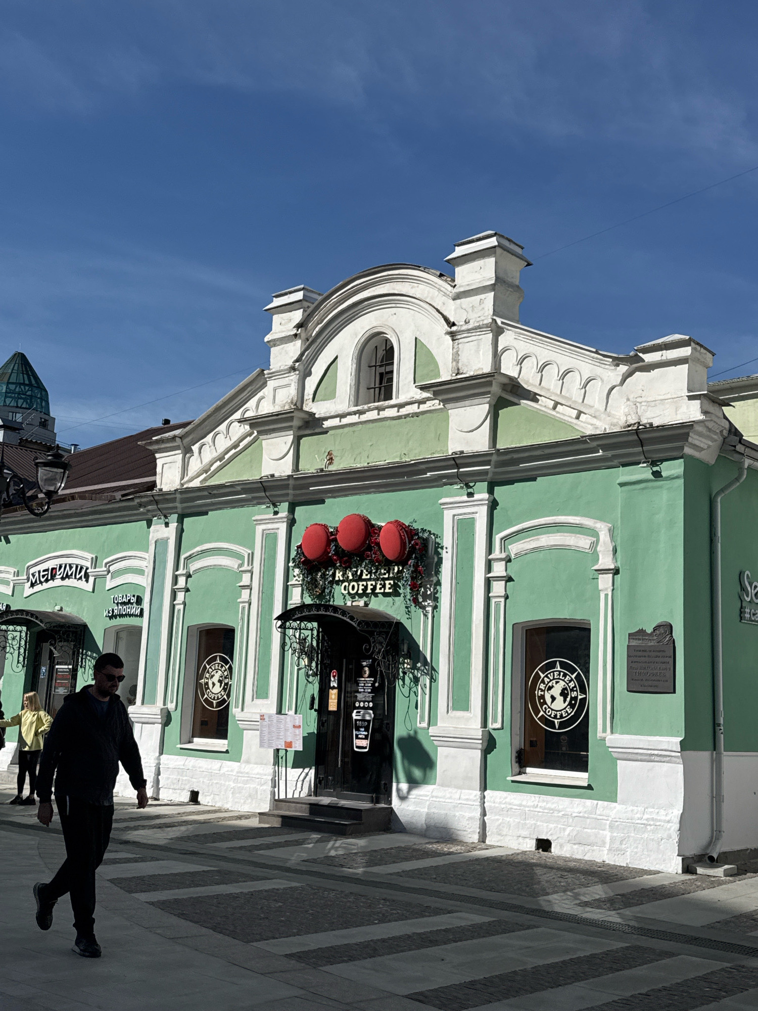 A man in dark clothing and sunglasses walks away from the camera on a paved sidewalk in front of a light green building. The building has white trim and features a large sign with red spheres and the words "TRAVELER'S COFFEE" in white. To the left of the coffee shop entrance, there is another sign in Russian that reads "МЕГУМИ" and below that "ТОВАРЫ ИЗ ЯПОНИИ" which translates to "JAPANESE GOODS". A woman in a yellow top and dark pants stands near the "МЕГУМИ" sign. The building is located on a sunny street with a blue sky overhead. The street in Tver, Russia, appears to be pedestrian-friendly with a patterned pavement.