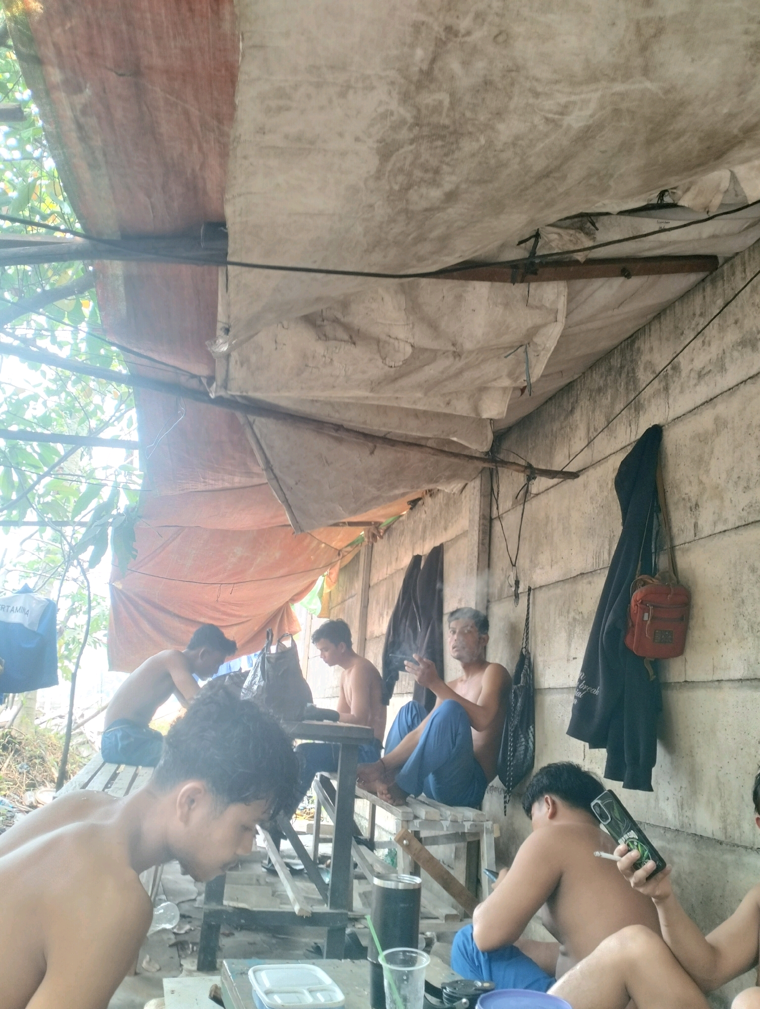 The image depicts a group of young men in a rustic, open-air shelter, likely in Pontianak, Indonesia. The shelter is constructed with a combination of tarpaulins and corrugated metal, providing partial cover from the elements. The environment appears to be somewhat cluttered, with debris and various objects scattered around the ground.
Four individuals are visible, all appearing to be young adults, with three of them topless and wearing blue pants. One man is seated on a wooden structure, actively smoking and holding a phone. Another man is similarly seated, looking down at something in his hands. A third man is partially visible in the foreground, also topless. A fourth individual is seated further back, also topless and wearing blue pants.
Several objects are present in the scene, including bags, a water bottle, a phone, and what appears to be a smoking apparatus. The overall atmosphere suggests a casual gathering or resting place. The wall in the background is made of concrete blocks, and clothes are hanging on it, including a dark jacket and a small red bag. The lighting indicates it is daytime, with sunlight filtering through the leaves of nearby trees.