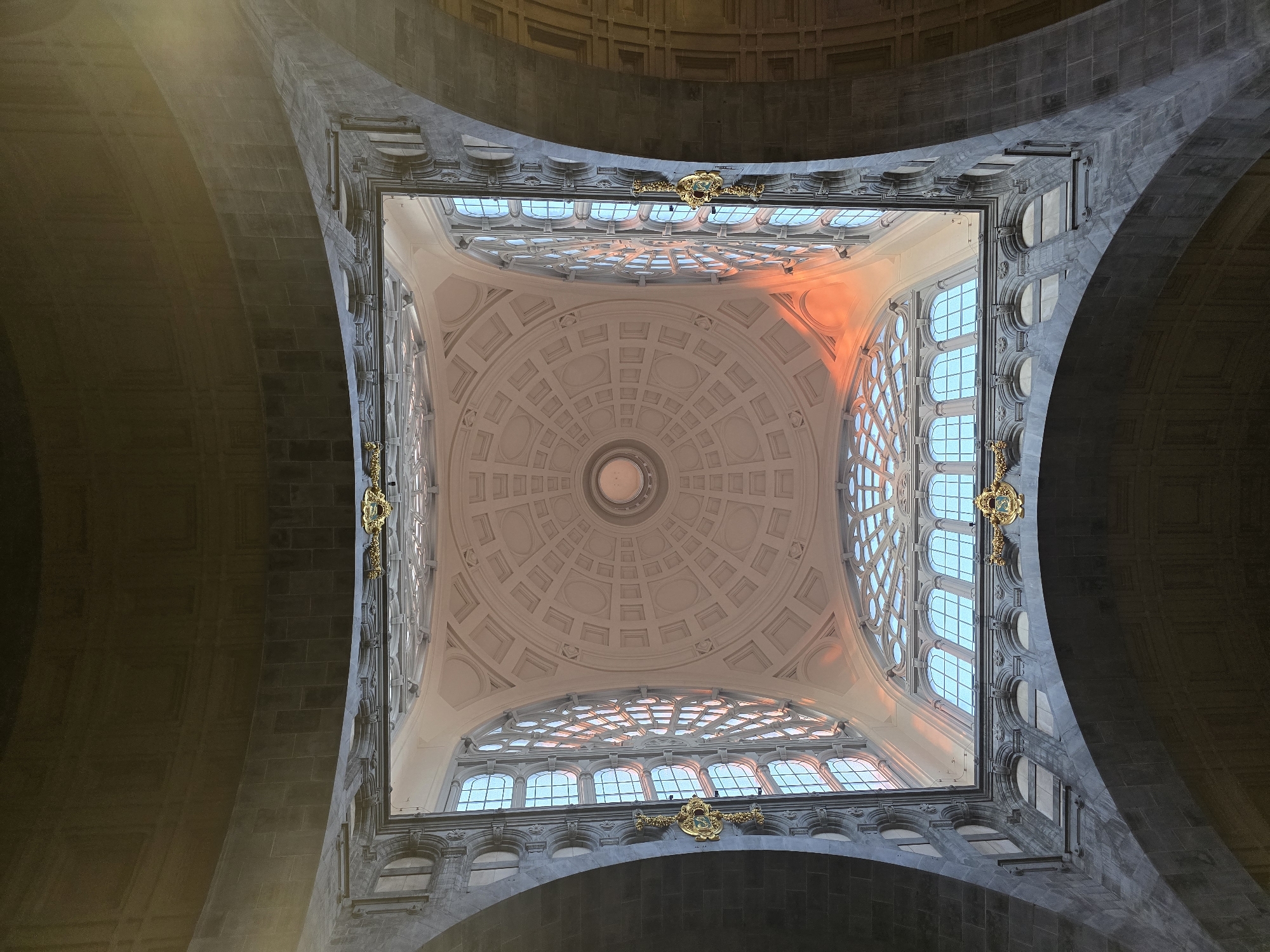 The image shows an interior view looking up at the domed ceiling of the Antwerp Central Station in Antwerp, Belgium. The dome is elaborate and features a circular opening at its apex, with concentric rings of architectural details. Large arched windows with intricate tracery surround the dome, allowing natural light to enter. The windows are segmented into many smaller panes, and the sunlight streaming through them casts a warm, orange glow on the ceiling and walls. The lower portion of the image shows the dark, stone-clad arches of the station's architecture. There are no people visible in the image.