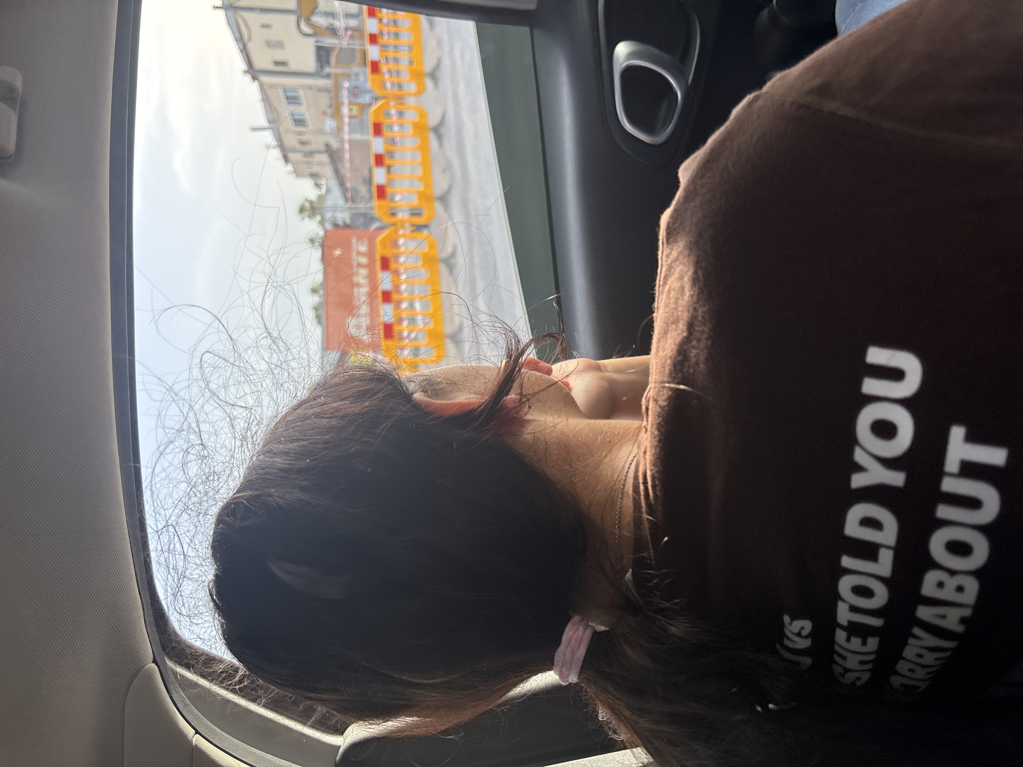 The image shows a young woman viewed from behind, seated in the back of a vehicle, looking out the window. She has long, dark brown hair pulled back in a ponytail with a light pink scrunchie. She is wearing a brown t-shirt with white text that reads "SHE TOLD YOU NOT TO WORRY ABOUT". Her left arm is resting on the window, and her hand is near her face. The background outside the car window appears to be a street scene with construction barriers and buildings, suggesting an urban or suburban environment, possibly in Urbanización Villa Martín, Spain, given the context. The lighting suggests it is daytime, with sunlight filtering through the window.