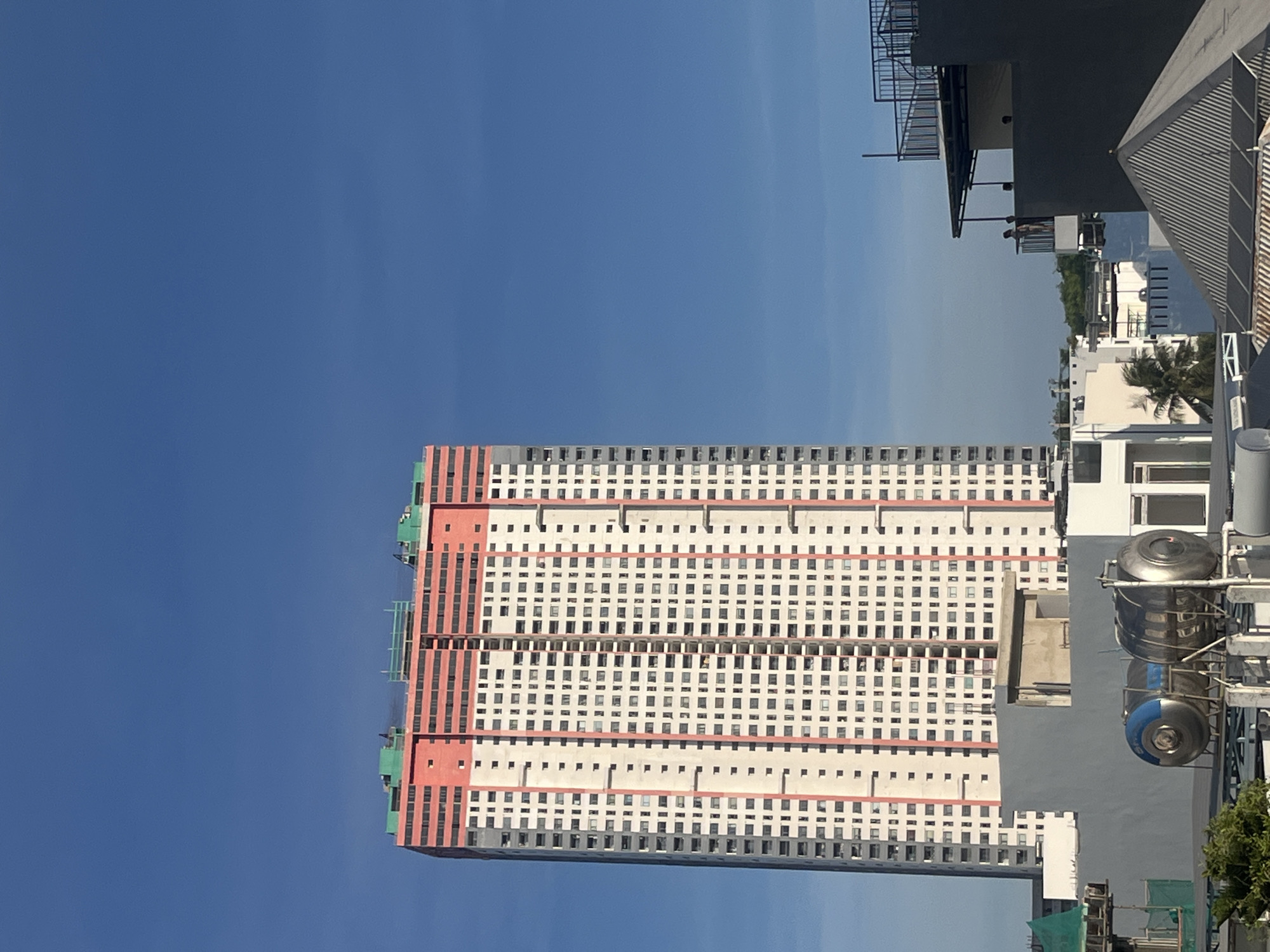 This is an outdoor, eye-level, medium shot of a large, modern apartment building in the city of Bac Nha Trang Ward, Vietnam. The building is tall and rectangular, with a white facade punctuated by rows of windows. The top portion of the building features a bright orange band and green structural elements, suggesting ongoing construction or recent completion. The sky is a clear, deep blue. In the foreground, several other buildings and structures are visible, including what appear to be water tanks on a rooftop and a multi-story building with a balcony where a person is standing. The overall scene depicts an urban landscape under a bright sky.