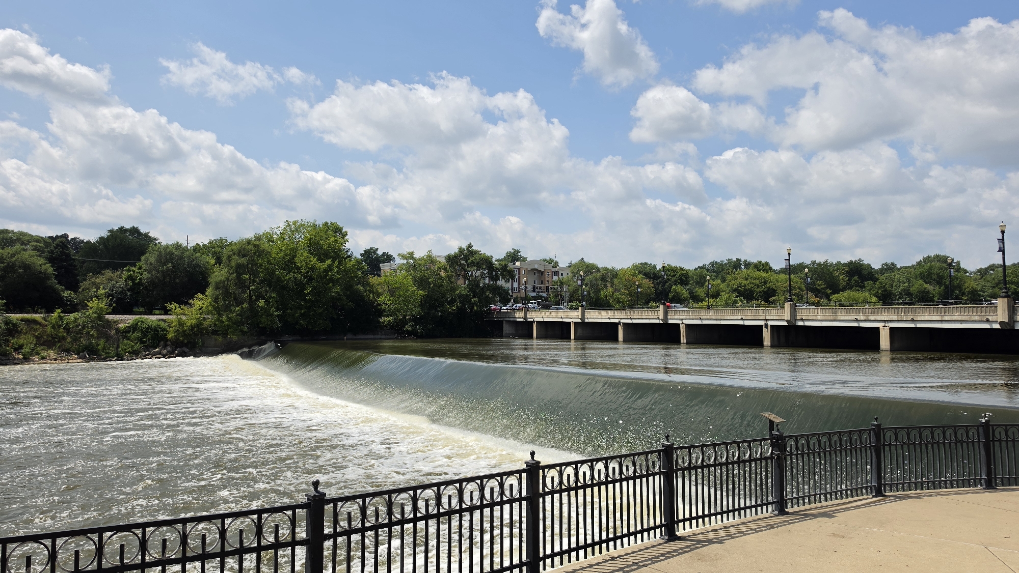 The image depicts a wide river with a man-made waterfall in the foreground, viewed from a paved walkway bordered by a decorative black metal fence. The waterfall cascades over a concrete dam, creating white, frothy water that flows into the calmer river. Beyond the waterfall, a long, multi-span bridge with concrete piers crosses the river. The bridge is lined with lampposts. On the far bank, a dense line of green trees forms the backdrop, with some buildings, including a multi-story tan building with windows, visible behind the trees. Cars are visible on the bridge and on the road behind the trees. The sky above is blue with scattered white, puffy clouds. The scene appears to be set in Elgin, Illinois, United States of America, given the context of the location. There are no people visible in the image. The time of day appears to be daytime, likely late morning or early afternoon, judging by the bright sunlight and shadows.