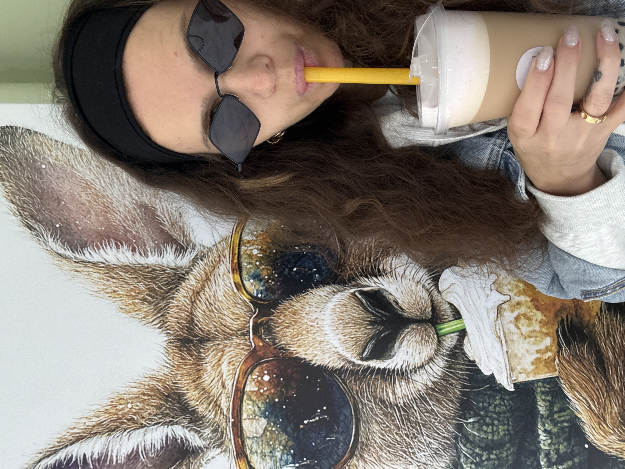 A woman is taking a selfie in front of a large portrait of a kangaroo wearing sunglasses and a scarf, with a drink in her hand. The kangaroo portrait is on a white wall. The woman has dark curly hair, is wearing sunglasses, a black headband, and a denim jacket over a light-colored hoodie. She is holding a plastic cup with a beverage and a yellow straw, which she is drinking from. The drink appears to be a light brown color with a foamy top layer. Her nails are painted with a glittery polish. She is wearing a gold ring on her left ring finger. There is a tattoo visible on her left hand. The background wall appears to be white, and there is a hint of a green wall in the upper right corner. The overall scene is casual and suggests the woman is at a cafe or a similarly relaxed establishment. There is no specific indication of its location in Moscow, Russia.