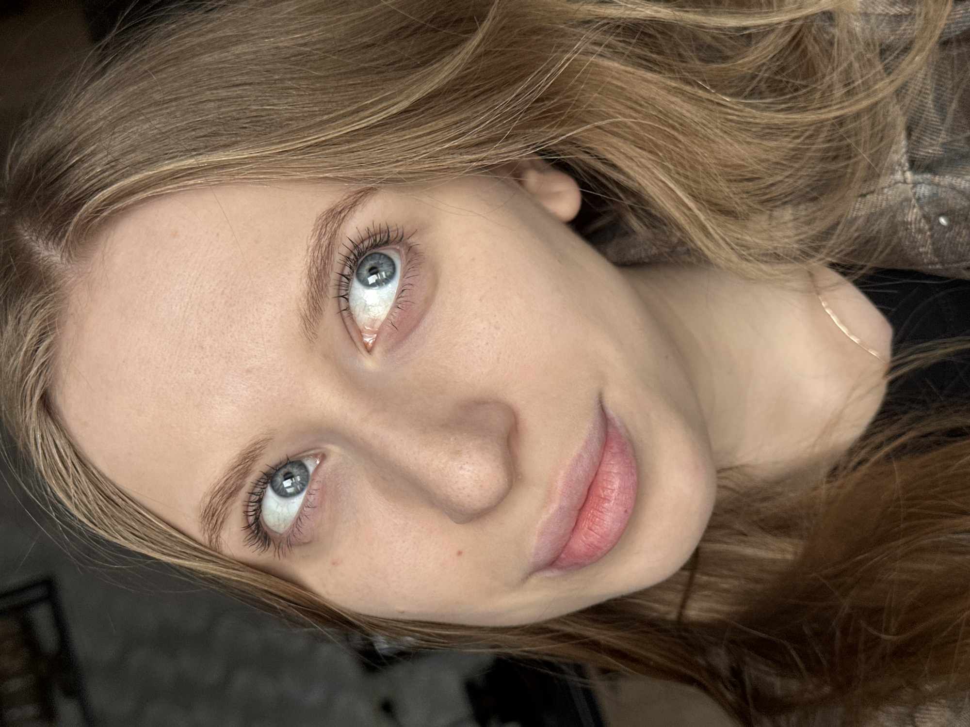 A close-up, angled portrait shows a young woman with fair skin, light brown hair, and light blue eyes looking up and to the left. Her lips are full and pink. Her hair is styled loosely around her face. A thin gold necklace is visible at her neck. Part of what appears to be a patterned garment is seen at the top right. The background is out of focus and indistinct. There is no discernible evidence to suggest a specific location, including Chaikovsky, Russia.