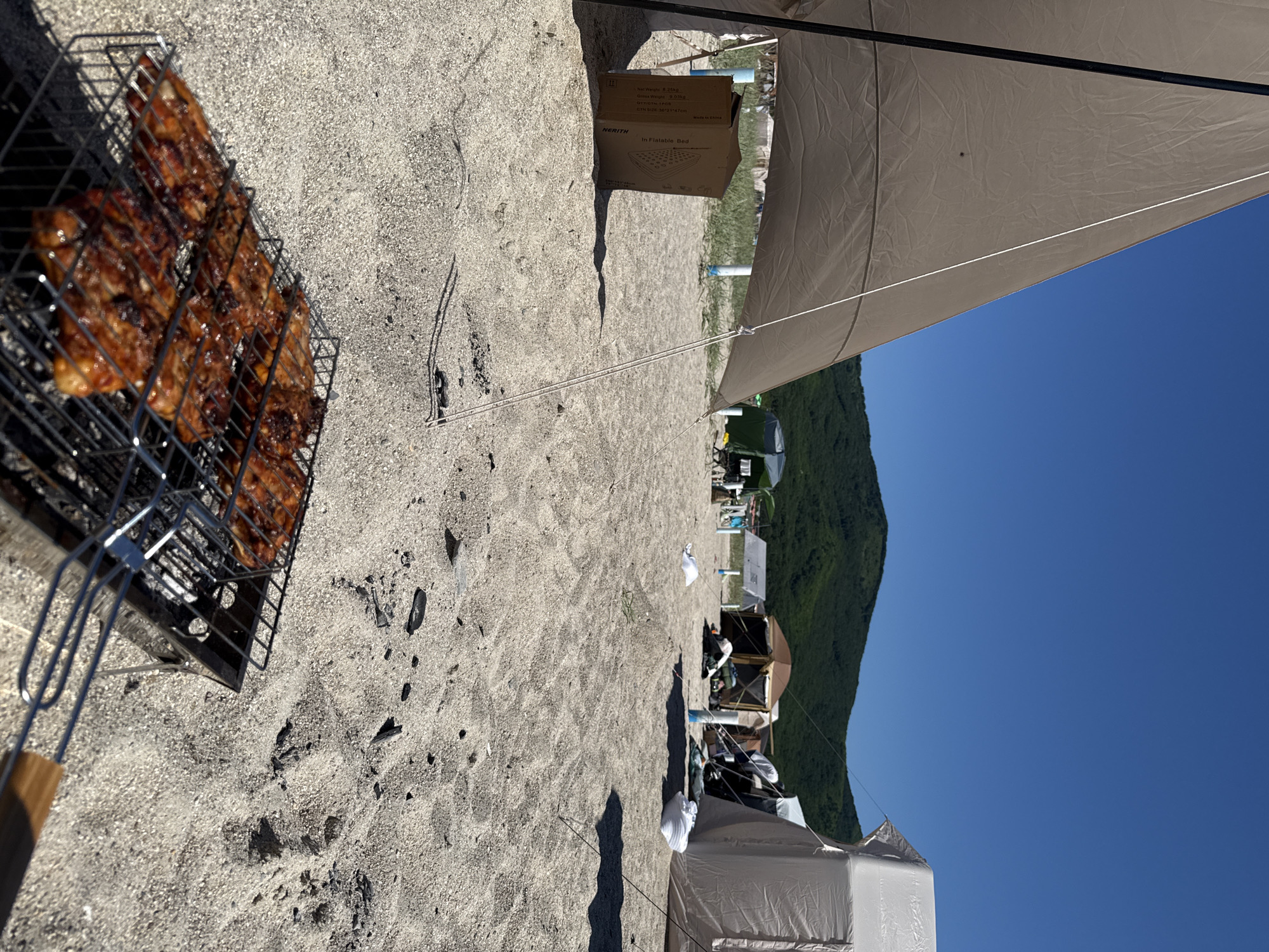 The image depicts a sunny day at a beach campsite, likely in the Nakhodka, Russia region, given the coastal setting with sandy terrain and a forested hill in the background. In the foreground, a charcoal grill holds several pieces of cooked meat, appearing to be chicken or pork, with visible grill marks and a glossy glaze. The grill is placed on a sandy surface scattered with small pebbles and dark debris, possibly burnt wood.
To the left of the grill, a large, tan tarp is extended from a pole, creating a shaded area. Partially under this tarp is a cardboard box, labeled "NERITY Inflatable Bed". Beyond this, several tents are visible, including a light-colored geodesic dome tent and a darker, more traditional tent. There are also some white objects scattered on the sand, which could be clothing or camping gear. In the far background, a lush green, tree-covered hill rises against a clear blue sky. The overall scene conveys a leisurely outdoor activity, possibly a barbecue at a beach campground.