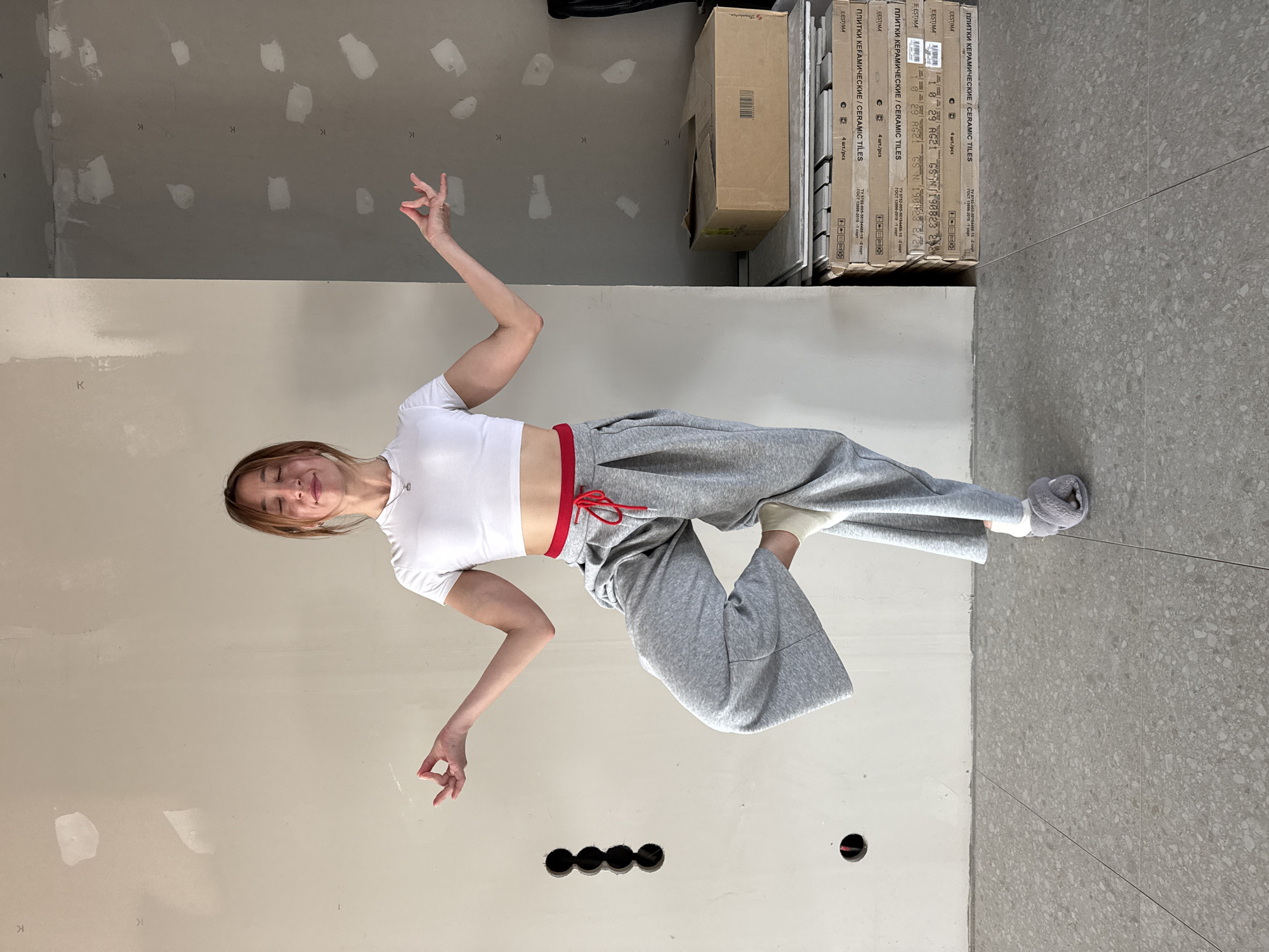 A woman is in what appears to be an unfinished room. The walls are a pale beige color, with the upper part of the walls unpainted and appearing as gray. The floor is tiled with a gray terrazzo pattern. The woman is wearing a white crop top, gray sweatpants with a red drawstring, and gray socks. She is standing on one foot, with the other foot lifted, in a dance-like pose. Her arms are outstretched. Behind her are stacks of boxes labeled in Russian, "CERAMIC TILES". The OCR indicates a location of "EESTIMA". This description is forensic, matter-of-fact, and contains only essential details. The city of Chaikovsky, Russia is not evident in this picture.