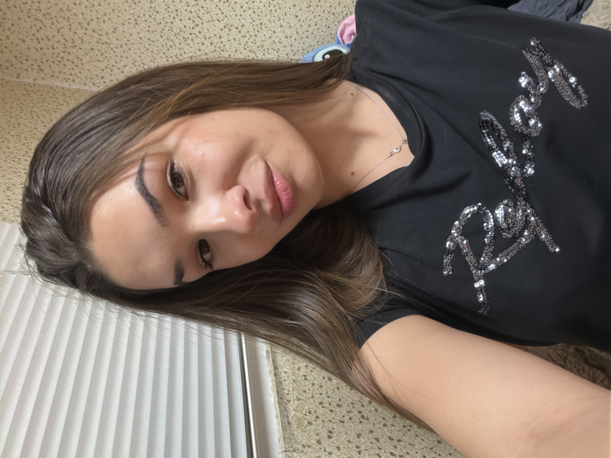 A young woman is captured in a close-up, tilted selfie, likely taken indoors in Sofia, Bulgaria. She has long, dark brown hair, neatly groomed dark eyebrows, and dark eyes, looking towards the camera with a soft, subtle smile and slightly parted lips. She is wearing a black t-shirt adorned with stylized silver sequined text across the chest. The first word of the text appears to be "BABYLON", followed by a second, less clearly legible word. A thin silver necklace with a small, elongated charm is visible around her neck, and a couple of dark moles are present on her upper chest/neck area. The background features a textured, light-colored surface on the right side and behind her head, and a white, vertically ribbed or pleated panel on the left. A blurry hint of colorful fabric, possibly a plush toy, is discernible near her head in the background. The lighting is soft and even, typical of an indoor setting, suggesting either daytime or evening.