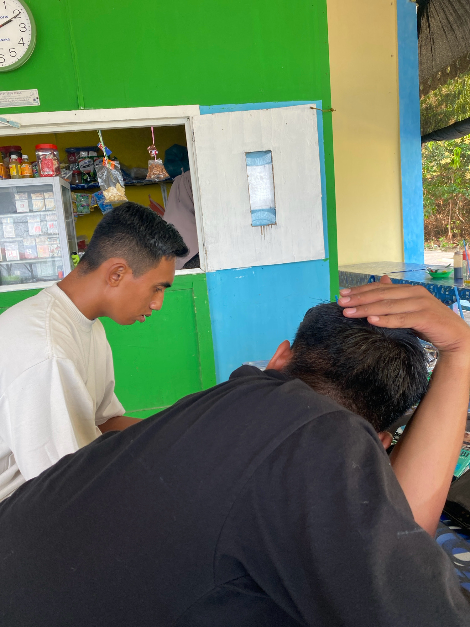 The image depicts two young men in what appears to be an outdoor food stall or small shop. The stall has a green and blue painted exterior. The man on the left, wearing a white t-shirt, is looking downwards with his mouth slightly open. The man on the right, wearing a black t-shirt, has his head tilted back and one hand resting on the back of his head, suggesting he may be resting or in a relaxed posture. Inside the stall, shelves are stocked with various items, including bottled drinks, packaged snacks, and possibly cigarettes. A clock on the green wall shows the time. In the background, trees and foliage are visible, indicating an outdoor setting. The overall scene is casual and appears to be in a warm climate. The presence of Indonesian text on a sign and the general aesthetic suggests the location is in Tanjung Pinang, Indonesia.