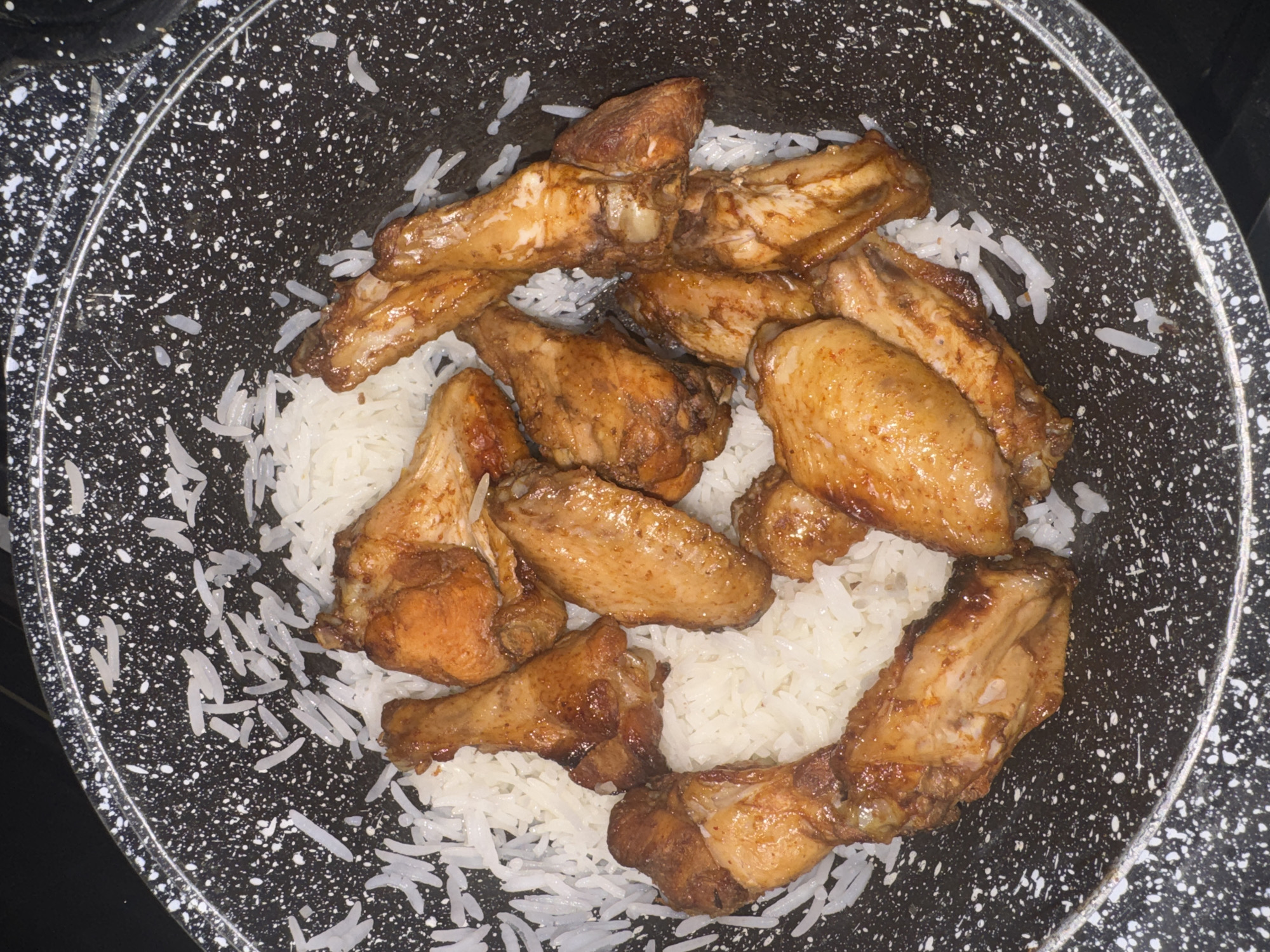 In a black speckled pot, a bed of white rice is topped with several golden-brown chicken wings. The wings appear glazed and possibly baked or fried. The pot's rim is visible, as is the dark background beyond it, suggesting the dish is likely indoors. There are no people or other objects of significance visible in the frame. The scene is a close-up, overhead shot of the food.
