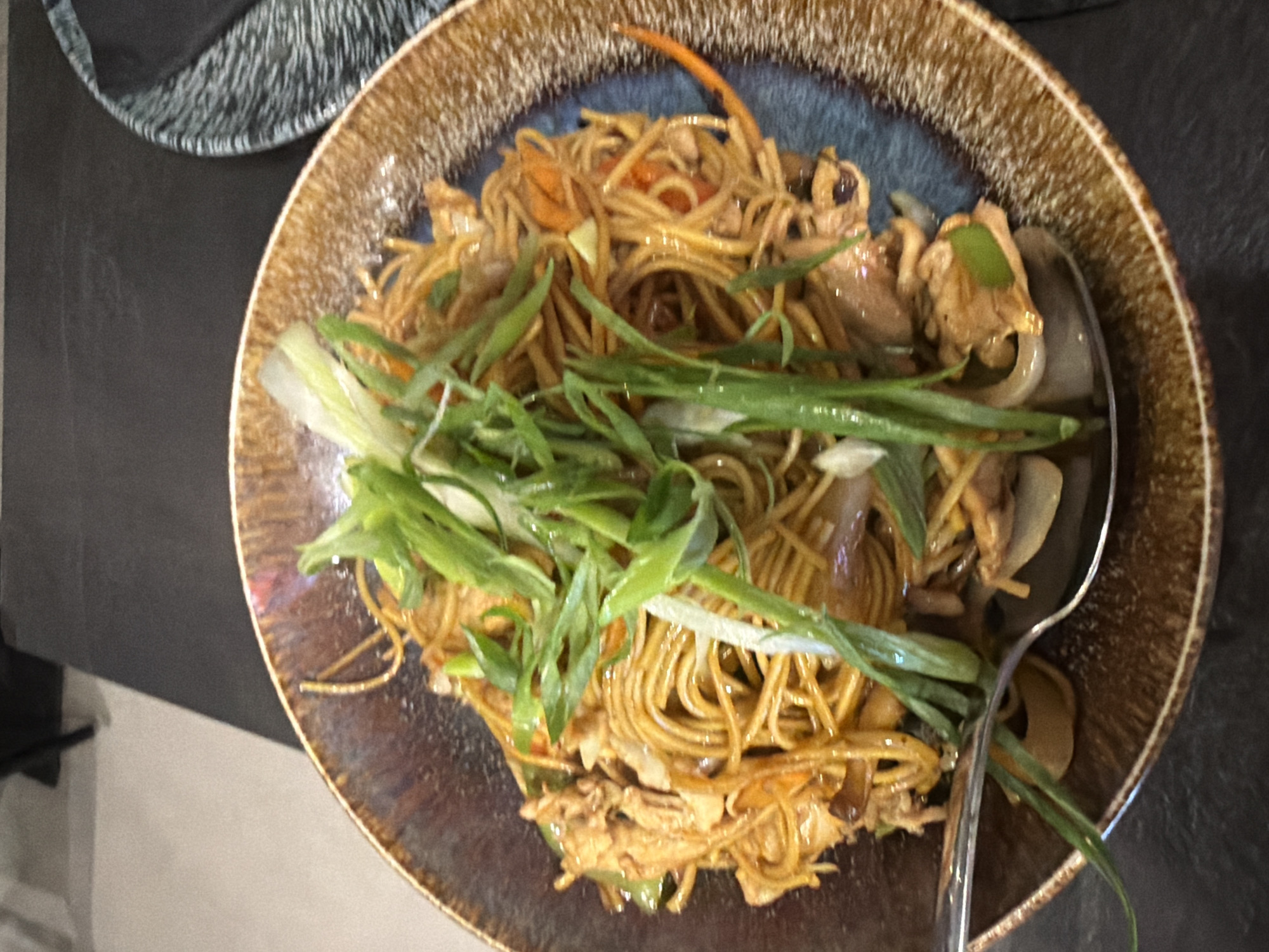 A close-up, overhead shot shows a bowl of noodles topped with chopped green onions, resting on a dark-colored table. The bowl has a rustic, textured brown and blue glaze. A silver spoon is visible in the lower left corner, partially submerged in the noodles. The background is out of focus, but suggests a restaurant setting. The food appears to be a stir-fried noodle dish with meat and vegetables. The location is Kiti, Cyprus.