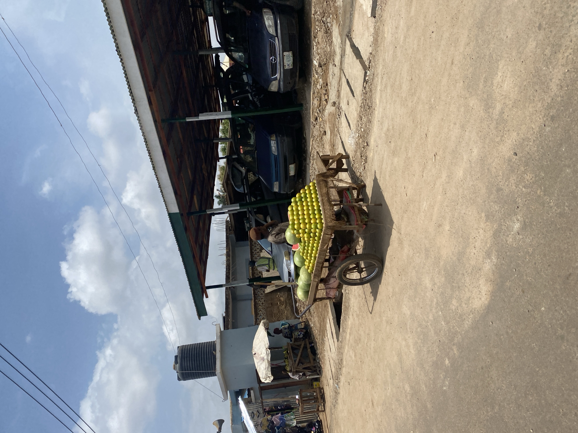 The image presents a vibrant outdoor scene, likely a market or street, in Kafanchan B, Nigeria, under a bright, partly cloudy sky. The central focus is a sturdy wooden cart, laden with produce, positioned on a light-colored, dusty road or concrete surface. Hundreds of small, green, spherical fruits, resembling limes or small oranges, are neatly stacked in rows on the cart, with a few larger green watermelons visible below them. A person is partially visible near the cart, presumably the vendor.
To the left of the cart, under a corrugated metal roof structure, several cars are parked. At least two dark-colored vehicles are discernible, with one clearly displaying the license plate "KMC 930DD". Further into the background on the left, a light-colored building is visible, topped with a black water tank. Another individual is seen standing under a white umbrella, possibly attending to an adjacent stall.
Power lines stretch across the upper left portion of the blue sky, which features scattered white clouds. The strong shadows cast by the cart and other elements indicate that it is midday or early afternoon. The scene captures a moment of everyday commerce and activity in an outdoor setting.