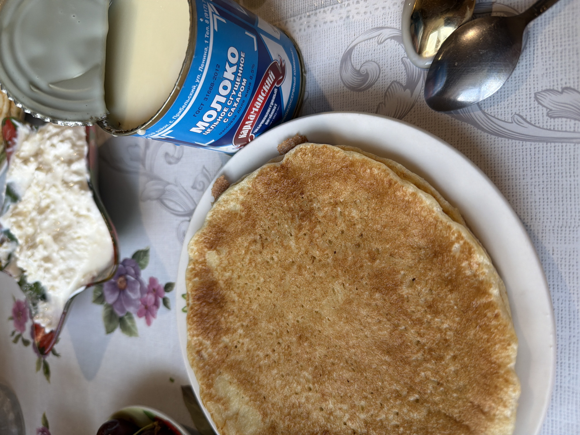 This overhead shot captures a home-style meal featuring traditional Russian elements. The scene is set on a table covered with a white tablecloth adorned with a subtle textured pattern and delicate purple and pink floral motifs.

The primary subjects are a stack of golden-brown pancakes or blini, presented on a simple white plate, and an open can of condensed milk. The condensed milk, labeled in Cyrillic, is partially visible and shows its creamy, light yellow contents. To the left, a portion of a creamer pitcher or small bowl filled with what appears to be cottage cheese or sour cream is visible, also decorated with floral designs that complement the tablecloth. In the upper right corner, two spoons, one reflective silver and one darker metal, rest on the tablecloth, indicating preparation for eating. A hint of another dish, possibly containing dark berries or jam, can be seen at the very bottom left.

The setting suggests an indoor daytime meal, likely breakfast or a snack, in a domestic environment. The items on the table indicate a sweet and comforting repast, common in Slavic cuisine.

Visible text on the condensed milk can identifies it as "МОЛОКО ЦЕЛЬНОЕ СГУЩЕННОЕ С САХАРОМ" (Whole Condensed Milk with Sugar) from the "Карламанский" (Karlamansky) milk cannery, with a fat content of "8.5%". An address on the label specifies "с. Прибельский, ул. Ленина, 1" (village Pribelsky, Lenina street, 1), and a GOST (State Standard) number "31688-2012" is also present, further confirming a Russian origin, consistent with the provided location context of Akkuzovo, Russia. The overall impression is one of a simple, comforting meal prepared and enjoyed in a Russian household.