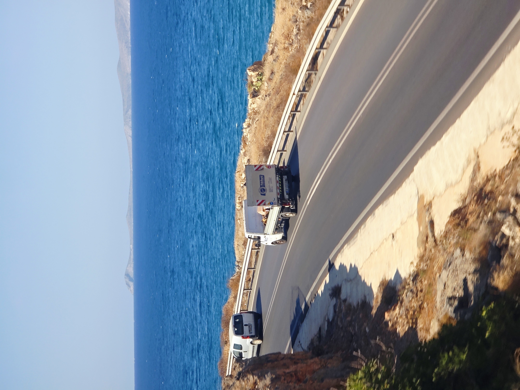 The image captures a coastal road in Gouves Municipal Unit, Greece, on a clear, sunny day. The road, asphalted and marked with double white lines, curves along the coastline. On the left side of the road, a white van is visible. On the right side, a white truck with a light-colored cargo box is traveling in the same direction. The truck appears to be carrying some boxes.
The sea is a vibrant, deep blue, stretching out to the horizon. In the distance, islands or peninsulas are faintly visible against the bright blue sky. The roadside is characterized by dry, arid vegetation, rocky outcrops, and a metal guardrail separating the road from the steep drop-off to the sea. The overall scene depicts a typical Mediterranean coastal landscape with active road traffic.