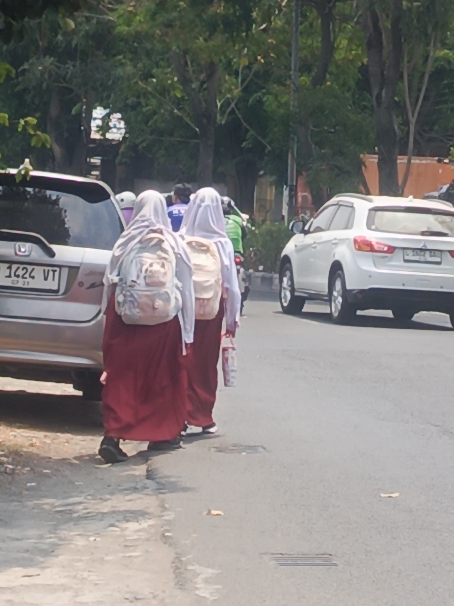 The image captures a street scene in Surabaya, Indonesia, under a bright sky. Two young individuals, dressed in school uniforms consisting of white headscarves and long maroon skirts, walk away from the camera. They are both wearing backpacks; one backpack is light-colored with a patterned design, and the other is a solid, lighter shade. The individual on the right is carrying a bottle in her hand.
They are walking on the side of a paved road. To their left, a silver SUV is parked. The SUV has a license plate that reads "B 1424 VT". Further down the road, a white SUV is visible, also on the side of the road. In the background, lush green trees line the street, suggesting a suburban or residential area within Surabaya. The overall lighting indicates it is daytime, likely late morning or afternoon, given the shadows.