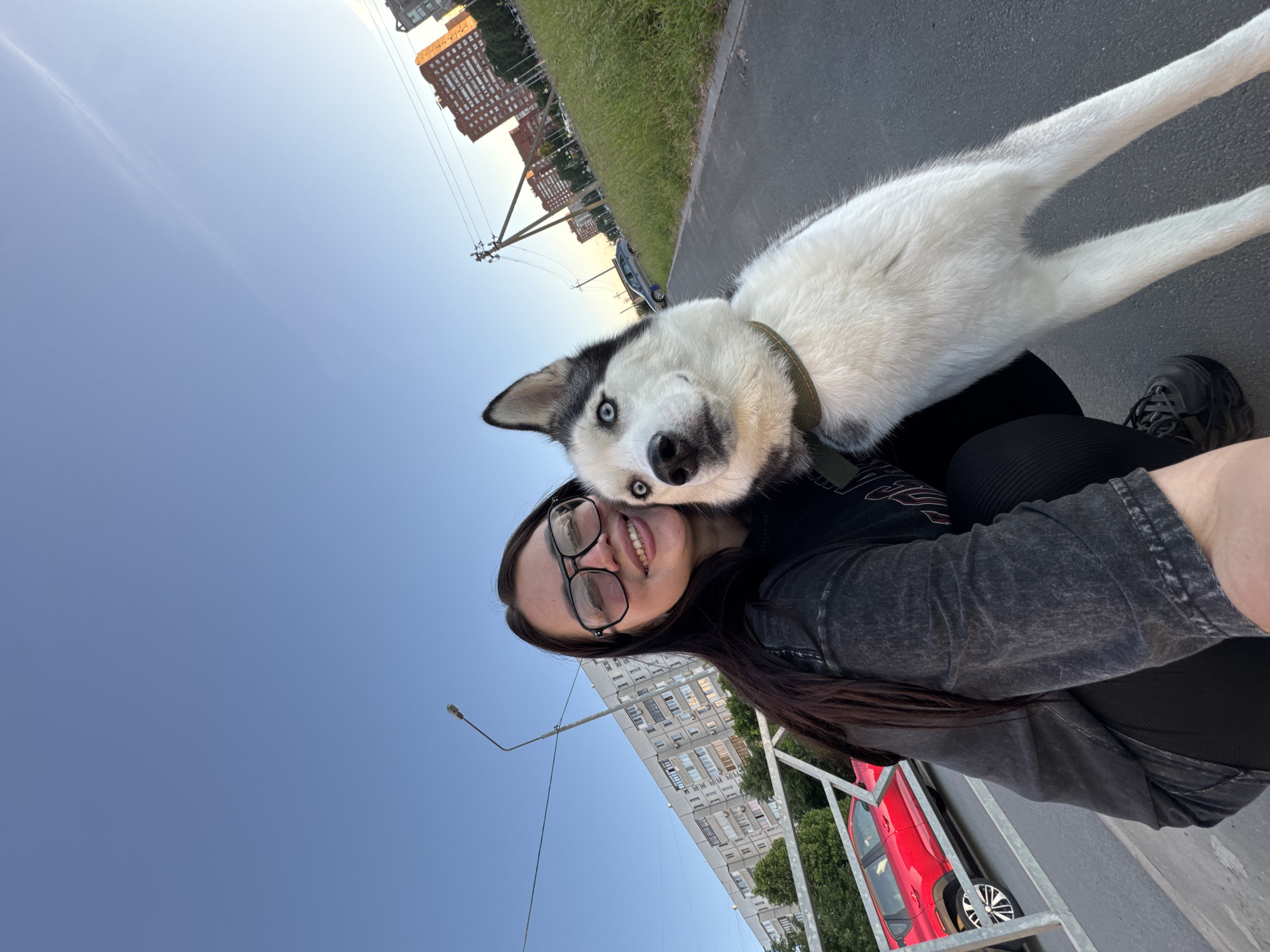 The image captures an upward-angled selfie-style shot of a young woman and a black and white Siberian Husky, likely taken outdoors during late afternoon or early evening. The photo is rotated, with the horizon appearing diagonal.
The woman is positioned on the right side of the frame, smiling widely at the camera. She has long, dark hair, is wearing black-rimmed glasses, a dark t-shirt with a subtle pattern, and dark pants or leggings. Her left arm and shoulder are visible, extending out of the frame.
A black and white Siberian Husky, with striking light blue eyes and a dark collar, rests its head on the woman's shoulder, looking towards the camera. The dog's white fur and partial body are visible towards the upper left.
The background shows an urban or suburban setting. Several multi-story residential buildings, some red-bricked and others lighter, are visible in the distance. A paved road or sidewalk runs along the ground, accompanied by green grass and utility poles with overhead wires. A street lamp extends into the clear blue sky, which occupies a significant portion of the upper left. A red car is partially visible, parked behind a metal railing on the lower left.
The lighting suggests a pleasant, clear day. The interaction between the woman and the dog conveys a sense of affection and happiness. No readable text is visible in the image.