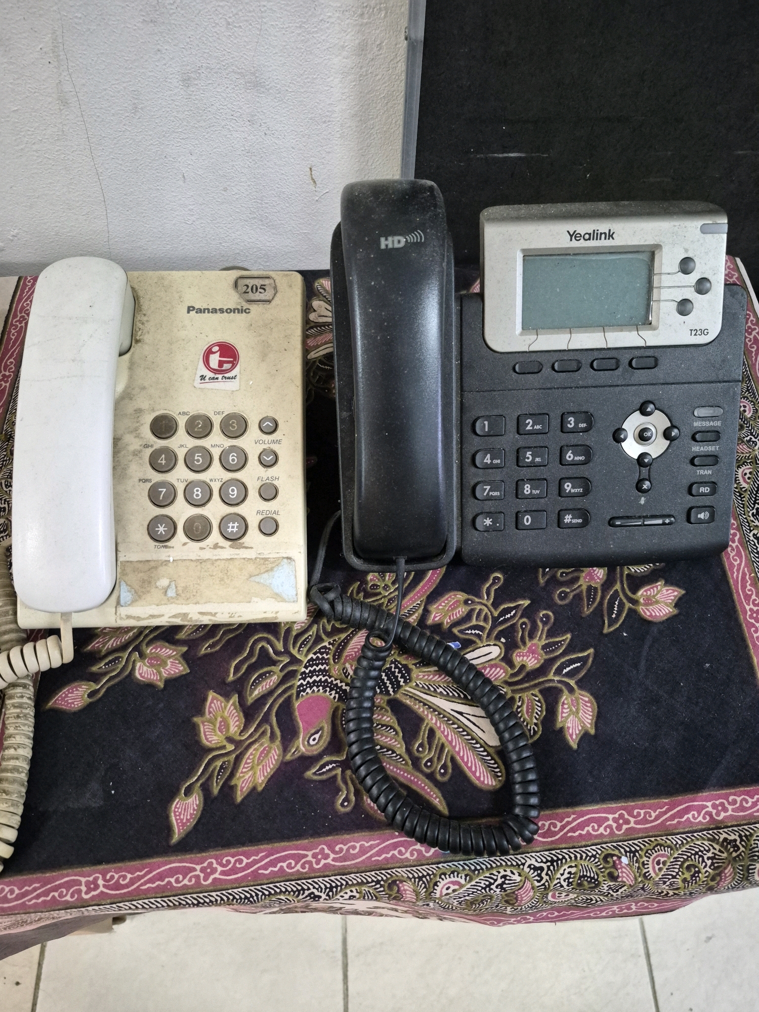 The image shows a top-down view of two telephones on a patterned fabric. The telephone on the left is a white Panasonic landline phone with a coiled handset cord. It has a keypad with numbers 0-9, *, #, and buttons labeled "VOLUME," "FLASH," and "REDIAL." A sticker with a red circular logo and the text "U can trust" is on the phone's body, along with "Panasonic" and the number "205" in a beige oval. The telephone on the right is a black Yealink T23G business phone with a digital display. It has a numerical keypad, directional buttons, and several function keys labeled "MESSAGE," "HEADSET," "TRAN," and "RD." A black coiled cord connects its handset. The phones are resting on a dark fabric with a pink and green floral and bird pattern. The background consists of a textured white wall and a dark surface. The scene is likely indoors, possibly in an office or a residential setting. This scene appears to be located in Purworejo, Indonesia, as indicated by the request.