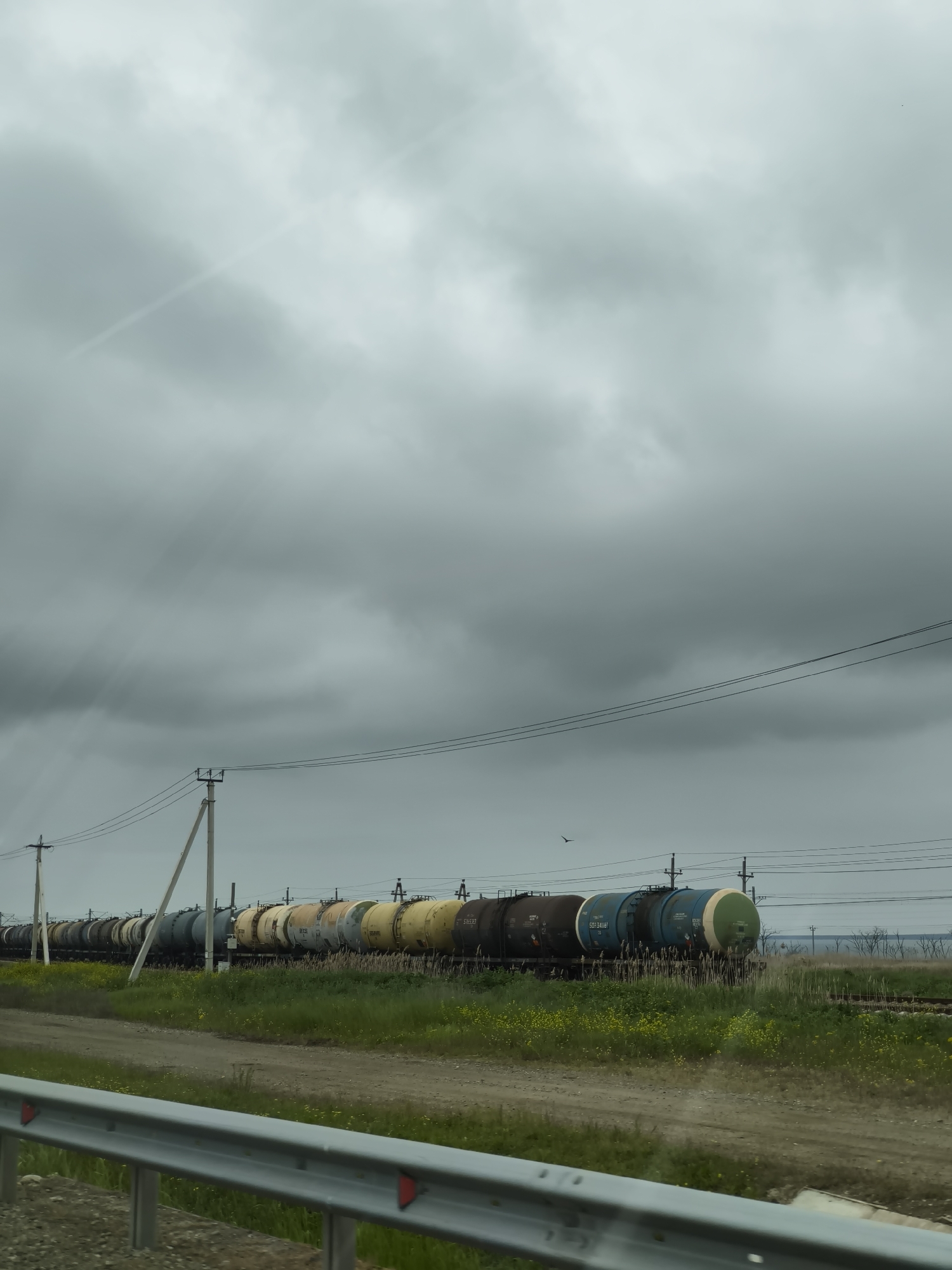 The image depicts a train of tank cars in the Ленинское сельское поселение, Ukraine. The tanks are of various colors, including yellow, brown, and blue. The train is situated on a track adjacent to a dirt road and green vegetation. Overhead power lines are visible above the train. The sky is overcast with heavy clouds. A metal guardrail is visible in the foreground.
