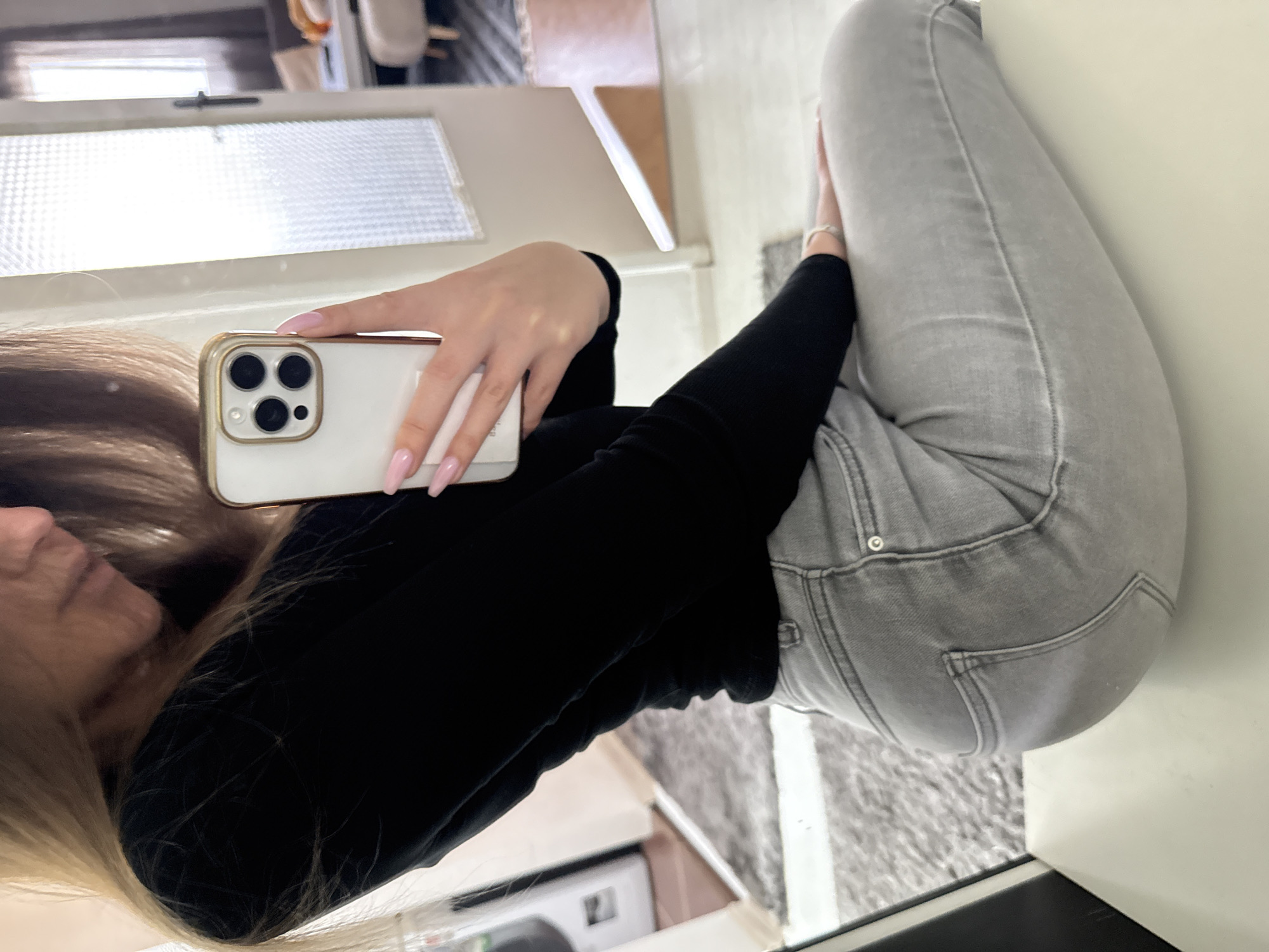 A fair-skinned person with long blonde hair and manicured nails is taking a selfie with a white iPhone inside a building. The person is wearing a black long-sleeved shirt and light grey jeans. The background shows a window and a glimpse of what appears to be a kitchen area with white cabinets. The exact location in Sofia, Bulgaria cannot be determined from the visual information provided.