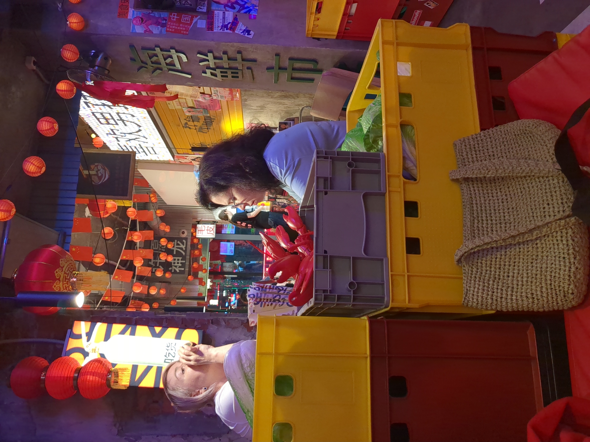 The image depicts an indoor scene in a restaurant or market stall in Tyumen, Russia, characterized by an East Asian theme. Two women are seated in the foreground and midground, surrounded by stacked plastic crates and various decorative and functional elements.
On the left, a fair-skinned woman with short blonde hair and a blue stud earring is wearing a white short-sleeved top. She is holding a small, dark object to her mouth, appearing to drink or eat. In front of her, partially obscured by yellow crates, is a large green leafy vegetable, possibly lettuce or cabbage.
In the midground, to the right of the blonde woman, a woman with dark, curly hair and glasses is seated, facing left. She is wearing a light blue short-sleeved top and has a neutral expression. Her right arm is slightly raised.
Between the two women, a stack of gray plastic crates contains multiple red, stylized plastic objects resembling lobsters or crawfish. To the right, another stack of yellow plastic crates contains green leafy vegetables. In the lower right foreground, a woven beige tote bag rests on a red cushion or mat.
The background features numerous red Chinese lanterns hanging from the ceiling and walls, along with string lights and neon signs. Prominent Chinese characters are visible on signs and walls, including a large vertical sign on the right reading "海鲜市" (Seafood Market) with "美食市" (Food Market) above it. Another illuminated horizontal sign reads "喜欢男" (Like men). Further in the background, smaller characters "神龙" (Divine Dragon) and "毛皮" (Fur) are visible. Walls are a mix of textured gray concrete and yellow corrugated paneling, adorned with various posters and flyers, one featuring a red anthropomorphic character. A black signboard with a bowl illustration hangs on the left. A ventilation fan is also visible. In the far background, a person in a dark t-shirt is partially visible, facing away.
