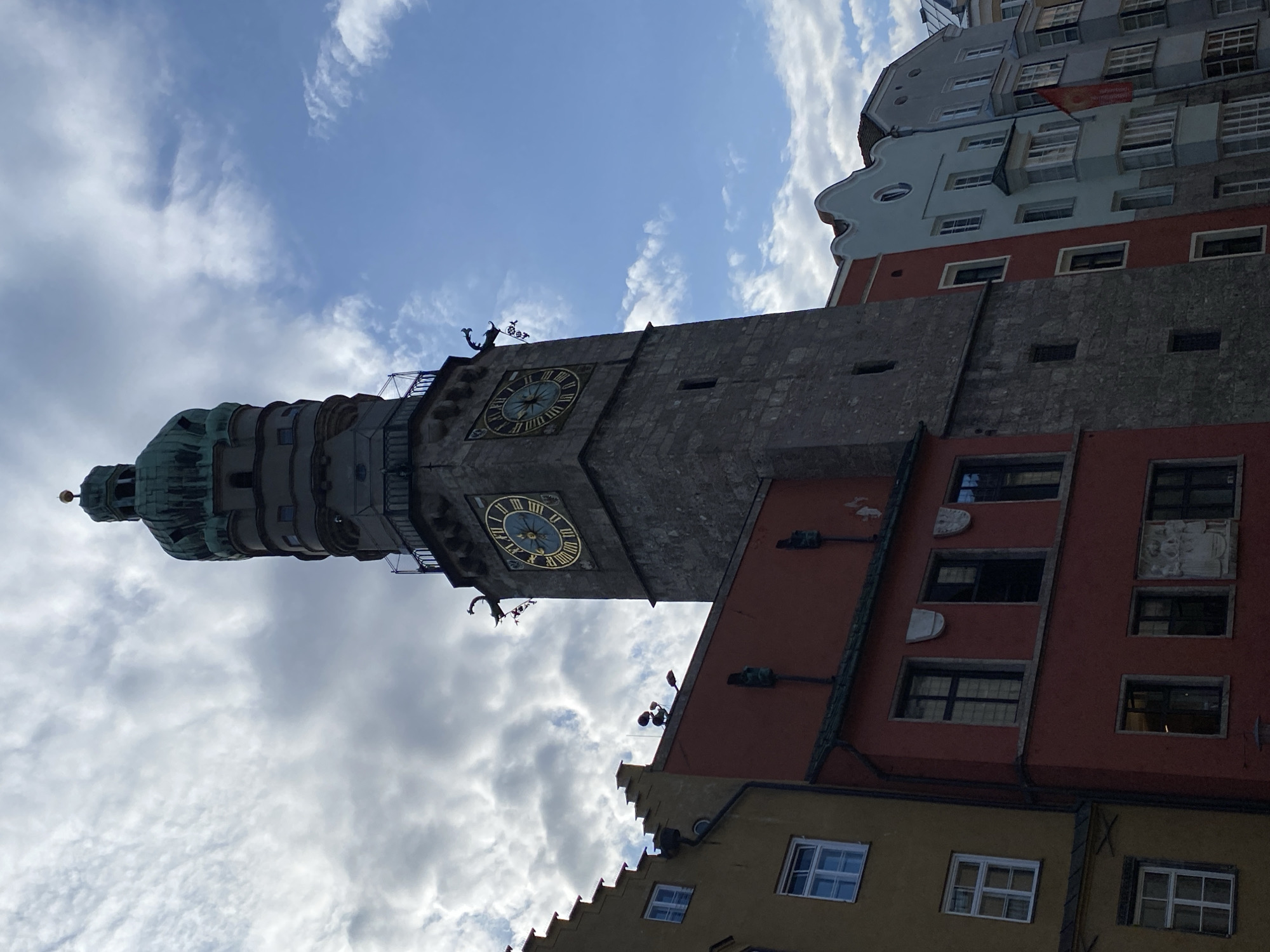 The image shows the Golden Roof (Goldenes Dachl) in Innsbruck, Austria, a landmark medieval building with a distinctive oriel window covered with golden tiles. The building is a tall, narrow structure with a stone tower topped by a green, onion-shaped dome. Two large clock faces are visible on the tower. Adjacent to the tower are several colorful buildings with distinct architectural styles, including a red building with coats of arms and a pale blue building with a unique gabled roofline. The sky above is partly cloudy, with patches of blue sky visible. The perspective is from ground level, looking up at the structures. There are no people visible in the image.