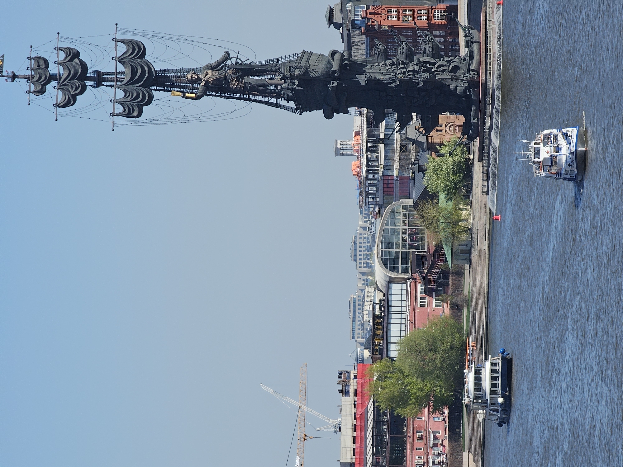 The image shows a vertical view of the Peter the Great statue in Moscow, Russia, along the Moskva River. The statue, depicting a ship with sails, stands tall against the blue sky. Alongside the riverbank are buildings with various architectural styles, ranging from modern glass structures to traditional red-brick buildings. Two boats are visible on the water, one moving toward the statue and the other docked along the lower part of the image. A construction crane is visible in the background.