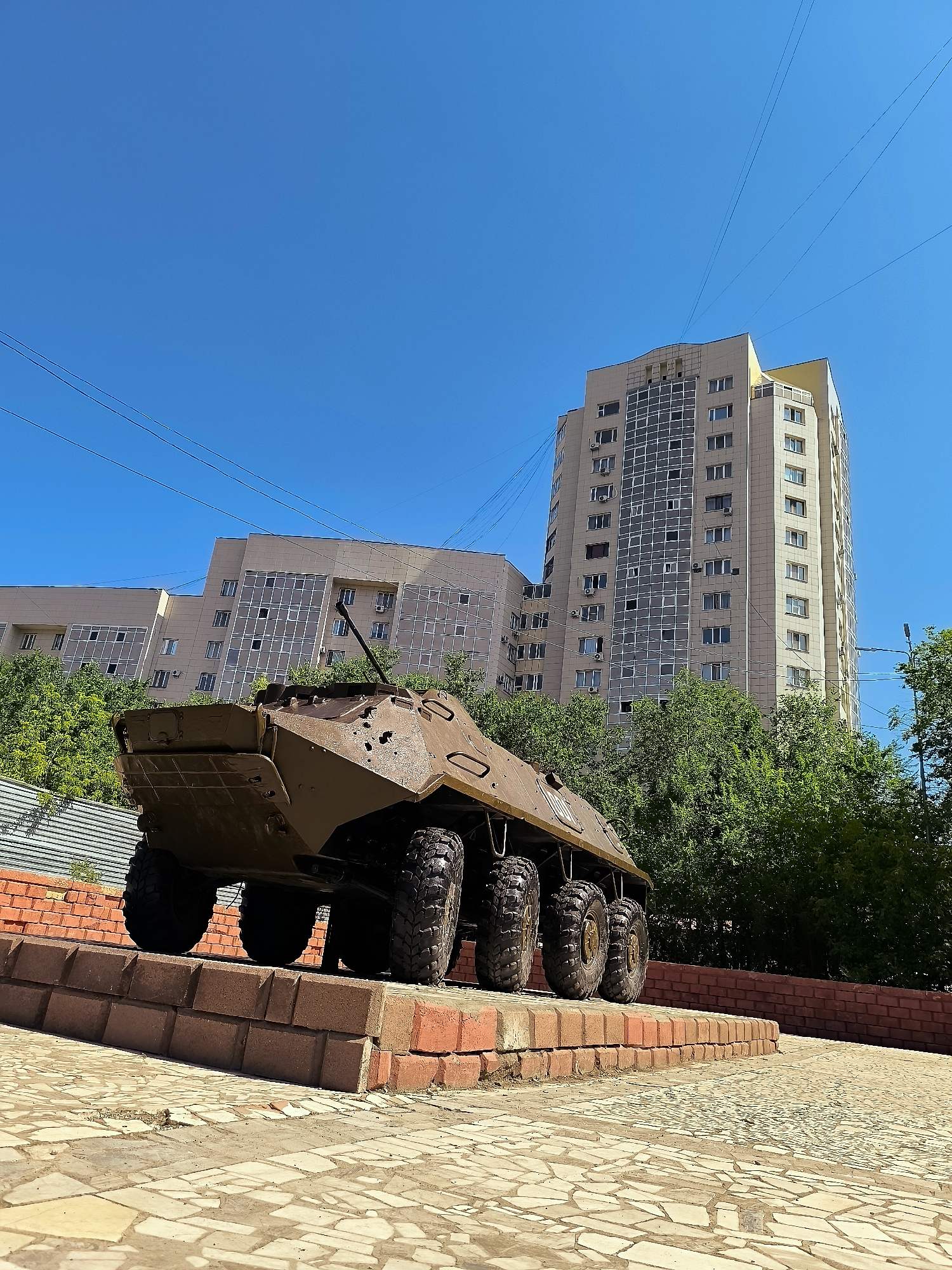 A brown armored personnel carrier is displayed on a brick pedestal outdoors, with a modern, multi-story building and trees in the background under a clear blue sky. The APC, positioned at a low angle perspective, faces away from the viewer, revealing its six large, treaded tires and the rear portion of its hull, which shows some signs of weathering and damage. The pedestal is constructed from red bricks and is situated on a paved area with a mosaic-like pattern. The building behind the APC is a tall, contemporary structure with numerous windows and balconies, suggesting an urban environment. Lush green trees partially obscure the lower sections of the building, indicating a green space within the city. Power lines are visible stretching across the sky.