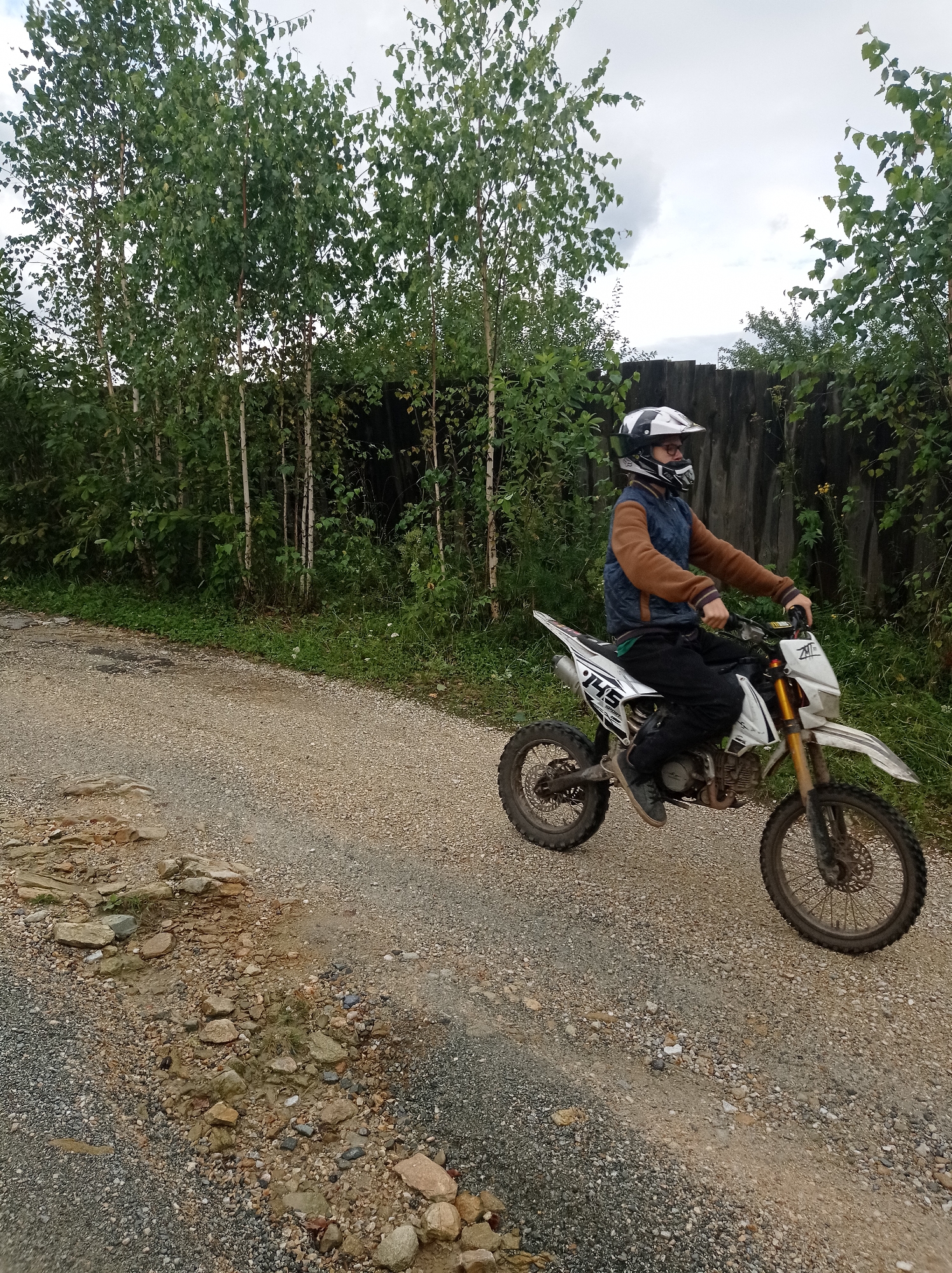 In Degtyarsk, Russia, a person wearing a helmet and goggles rides a dirt bike on a gravel path lined by trees and a wooden fence. The person is wearing a brown and blue jacket, black pants, and dark shoes. The dirt bike is white with green and black accents, and has the number "145" on the side. The sky is overcast. The terrain is uneven, with some rocks and puddles on the path.