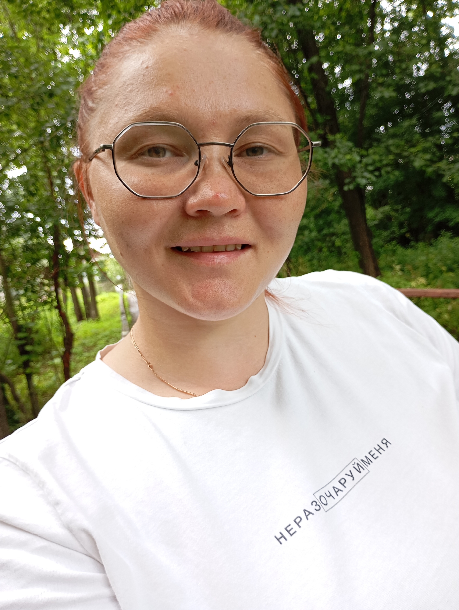 A young woman with reddish-brown hair pulled back and wearing clear, hexagonal-framed glasses is photographed in a selfie outdoors. She has fair skin with freckles and is wearing a white t-shirt with text in Russian on the right side. The text appears to be "НЕРАЗОЧАРУЙМЕНЯ" inside a rectangle. She is smiling faintly. The background is blurred, showing green foliage and trees, suggesting a park or wooded area. The lighting is natural and diffused. There are no visible landmarks that would specifically place this in Chaikovsky, Russia, beyond the assumed context.