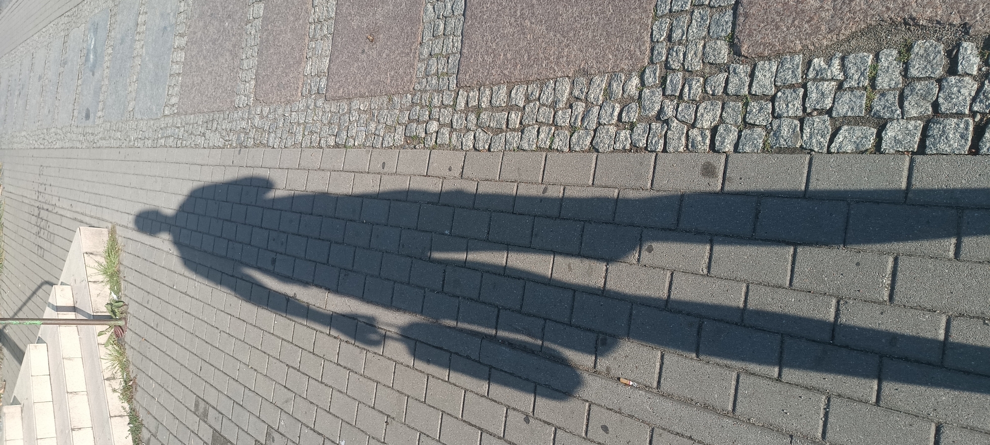 The image shows the shadow of a person on a paved sidewalk in Świnoujście, Poland. The shadow is cast by a person holding a bag in their left hand and standing with their body facing slightly to the right of the camera. The person's shadow extends from their head, which is visible near the top left of the frame, down to their feet, which are towards the bottom center. The shadow is long, indicating it was taken during a time of day with a low sun angle, likely morning or late afternoon.
The sidewalk is made of rectangular gray pavers, with a section of cobblestones and larger, darker stone slabs to the right. There are some stairs visible in the upper left corner of the image, adjacent to a metal pole that supports the shadow. Some sparse green vegetation is growing around the base of the pole and between the pavers. The overall scene depicts a common urban environment.