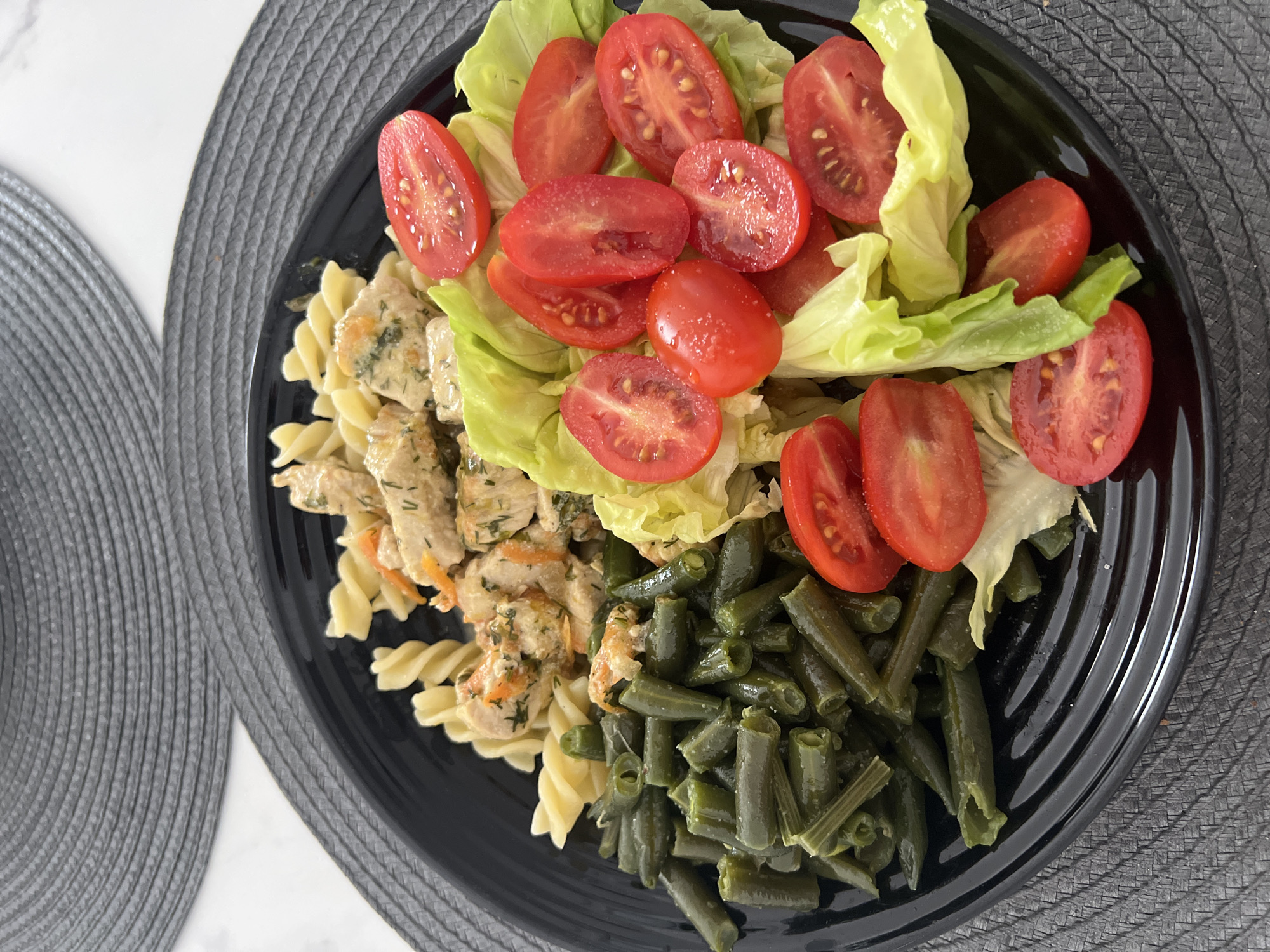 A black plate on a grey placemat holds a meal consisting of pasta, chicken with a creamy sauce, green beans, lettuce and sliced tomatoes. The scene is indoors. The location, Budva, Montenegro, is not discernible from the image.