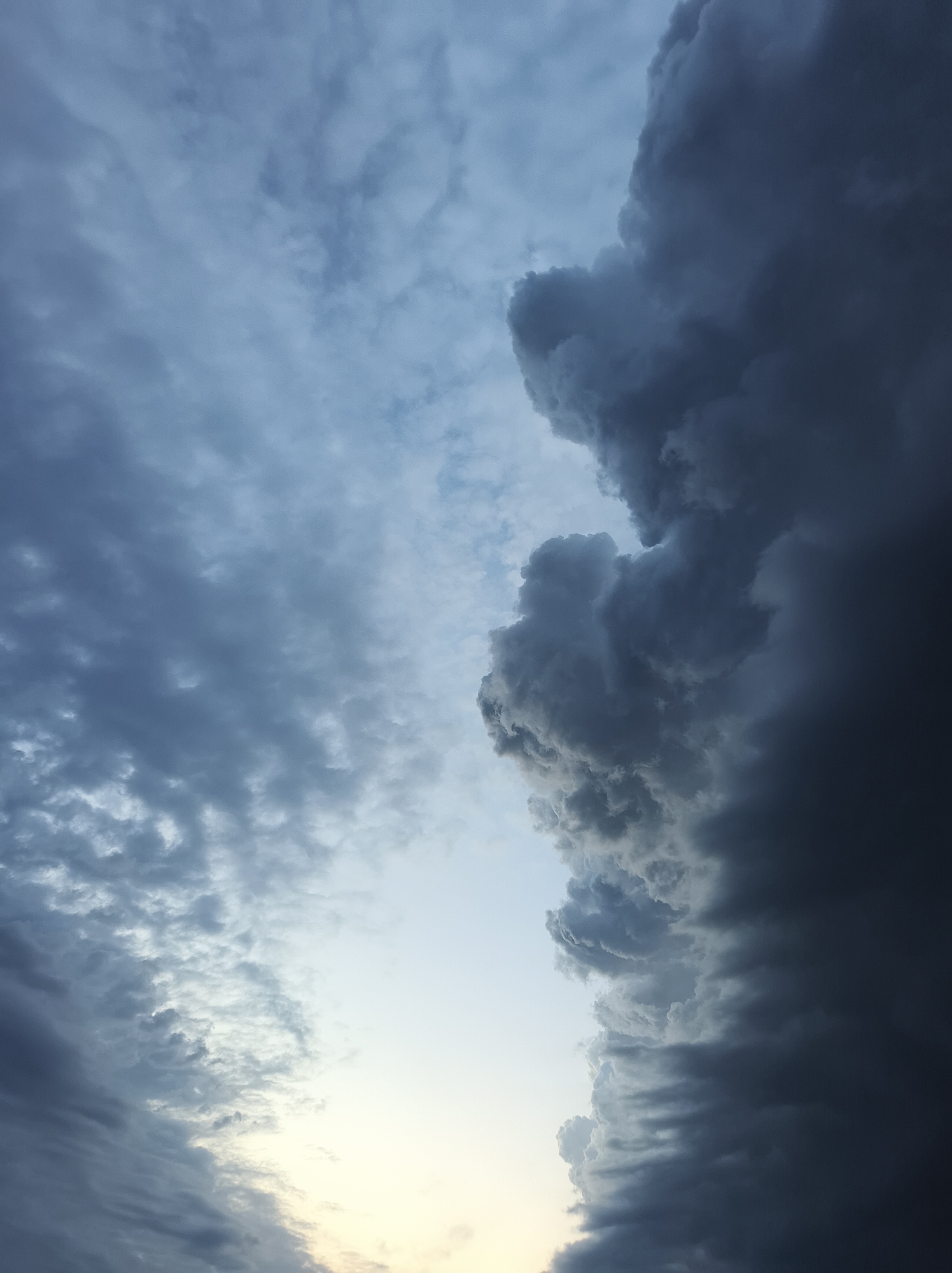 The image is a low-angle shot of a cloudy sky, likely taken outdoors. On the right side of the frame, a large mass of dark, billowing clouds dominates, appearing ominous and dense. In contrast, the left side displays thinner, lighter-colored clouds, creating a visual contrast. A small sliver of light, possibly from the sun, is visible at the bottom center, suggesting either sunrise or sunset. The image was taken in Vyshevychi, Ukraine.