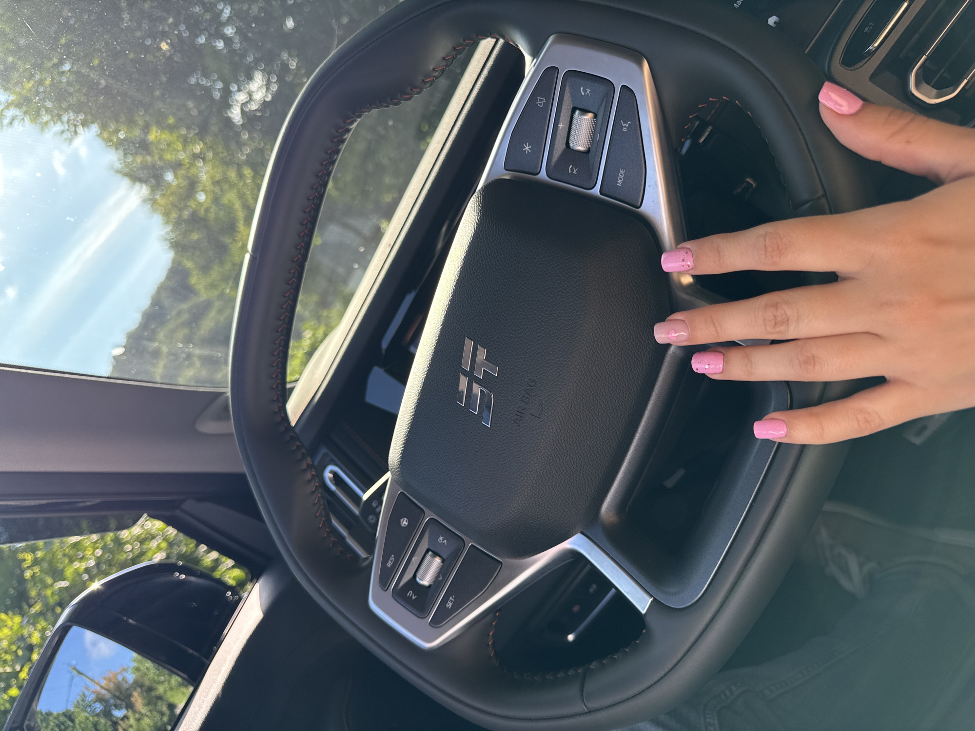 The image is a close-up, eye-level shot from inside a vehicle, focusing on the steering wheel and a person's hand with pink nail polish. The steering wheel is black leather with red stitching and silver accents. It features several buttons for vehicle controls, including "RES+", "SET-", "AV", and "MODE". The car's logo, a stylized "ST", is visible in the center of the steering wheel, along with "AIR BAG". The background shows a sunlit outdoor scene with green trees and blue sky, indicating it is daytime. The car's side mirror, reflecting trees and sky, is visible on the left. The person's hand, with manicured pink nails, is positioned over the lower part of the steering wheel, suggesting they are about to drive or have just finished driving. The overall impression is one of being inside a modern car on a clear, sunny day. There is no specific information in the image to place it in Kroyanskoye, Russia.