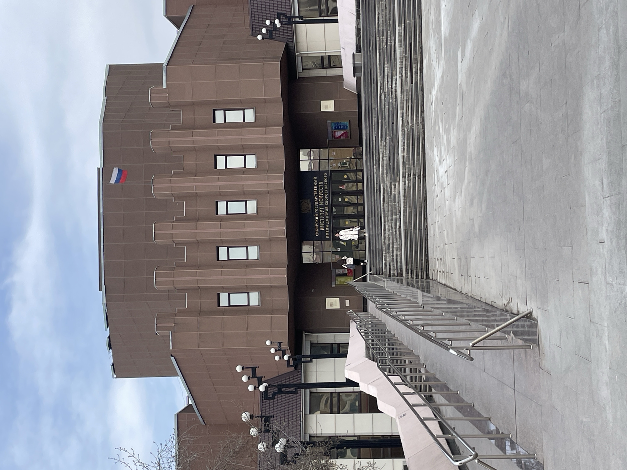 The image shows the Siberian State Institute of Arts named after Dmitry Hvorostovsky, located in the city of Krasnoyarsk, Russia. The building is a large, multi-story structure with a brown tiled facade, featuring a Russian flag displayed near the top. The entrance area is visible with a sign in Russian. A wide set of stairs with metal railings leads up to the entrance. Pedestrians are visible near the entrance. The sky is partly cloudy.