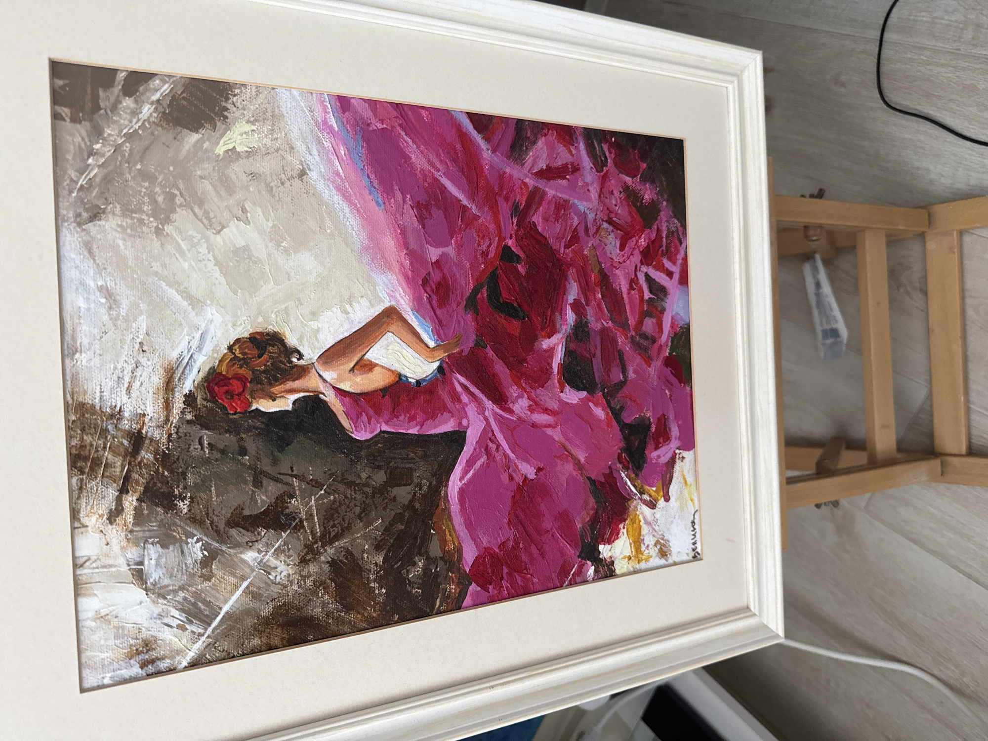 An oil painting on canvas, framed in white, depicts a flamenco dancer in a vibrant pink dress. The dancer, with a red flower in her hair, is captured mid-pose, her skirt dramatically swirling around her. The background is abstract, rendered in earthy tones of brown, gray, and white, with visible brushstrokes suggesting movement and texture. The painting is displayed on a wooden easel. The floor beneath the easel appears to be light-colored wood. The scene is indoors, with a hint of white molding visible on the left side, possibly indicating a wall. There is a white cable running across the floor from the lower right. The image does not provide any information about its location in Burgas, Bulgaria.