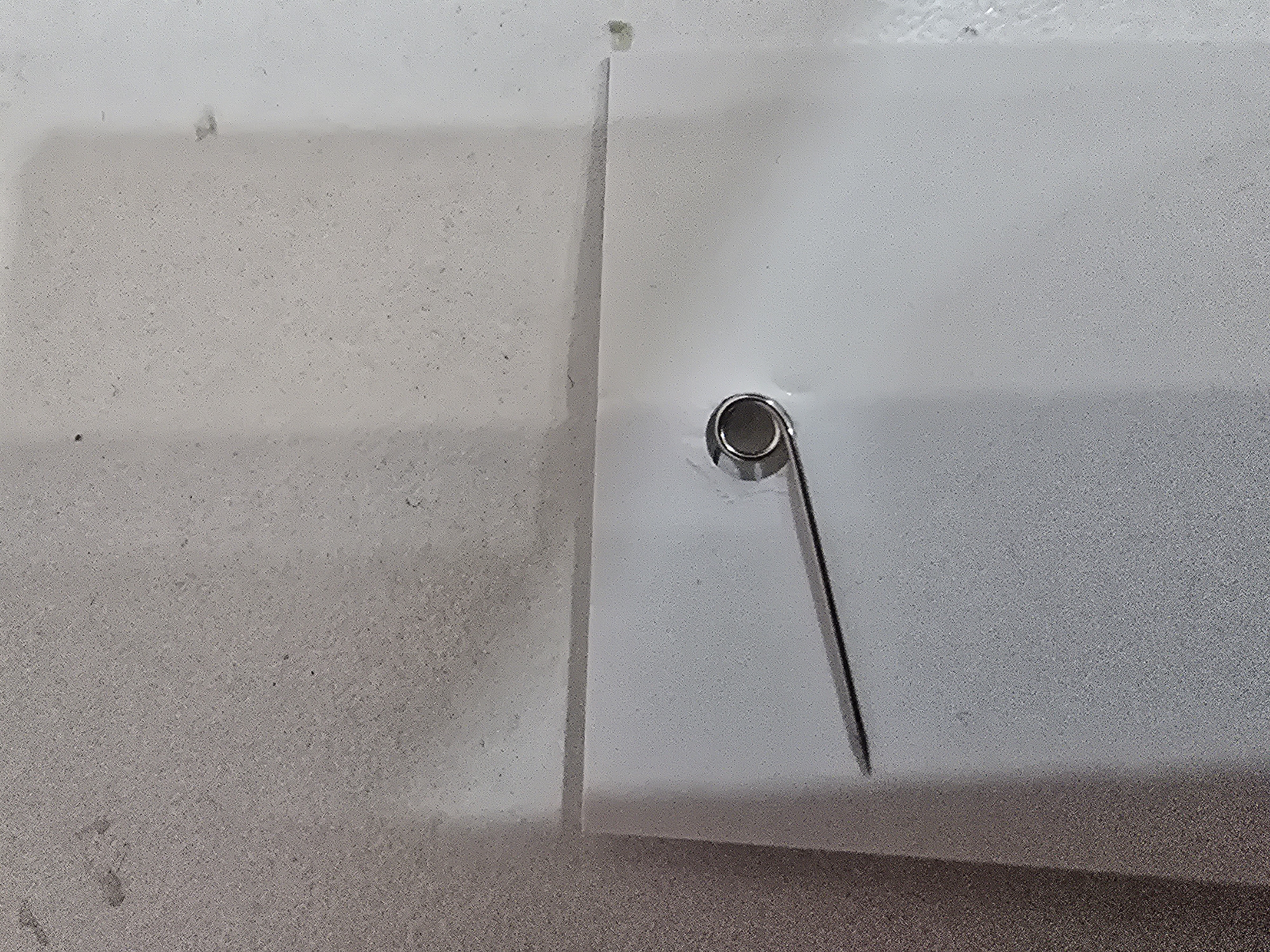 The image shows a close-up of a white surface with a thin, dark object extending from a silver, coiled spring. The surface appears to be a flat, white panel, possibly part of a larger object or structure. The coiled spring is metallic and circular, with the dark object inserted into its center. The dark object is thin and appears to be made of metal, extending horizontally across the white surface. The background is textured and appears to be a grey, rough surface. The lighting in the image is uneven, with some areas being brighter than others, casting shadows. The image does not contain any elements that would suggest a specific location within the city of Ulan-Ude, Russia, or any other identifiable urban environment. The scene is sterile and devoid of context beyond the objects themselves.