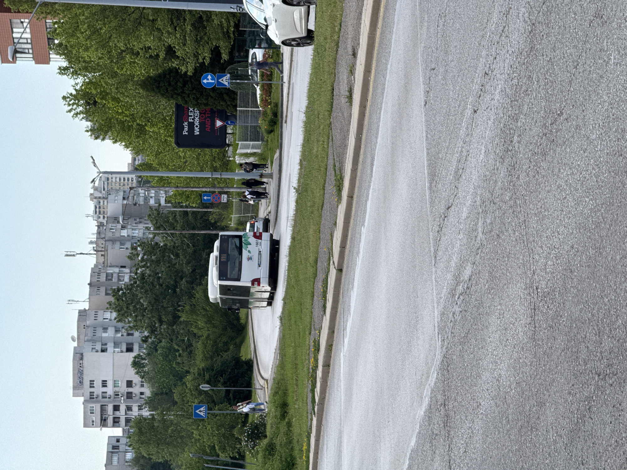 The image shows a street scene in Sofia, Bulgaria. A white MAN Lion's City bus with the license plate CB 1697 is driving on the right side of the street. A black billboard with the words "Park Share Flex Workshop To Go And The" is visible. Pedestrians are walking along the sidewalk. In the background, there are residential buildings and trees. A white car is parked on the right side of the street in the distance. Traffic signs are present.