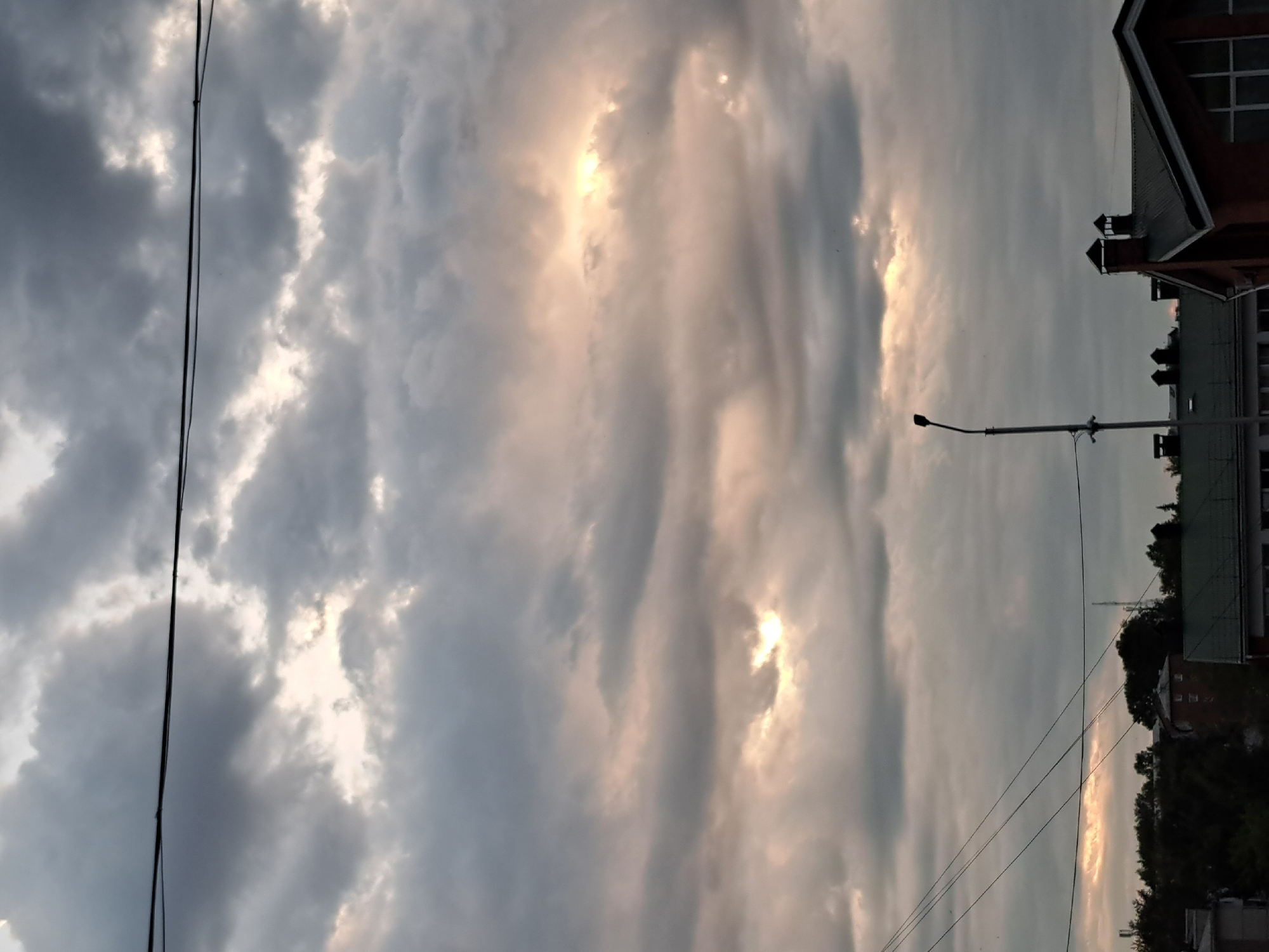 The image captures a cloudy sky over a city, possibly Irkutsk, Russia. In the foreground, a streetlight stands on the left, casting a long shadow. To the right, the rooflines of several buildings are visible, including one with prominent chimneys. The sky is filled with dramatic clouds, with patches of sunlight breaking through, illuminating the scene with a soft, ethereal glow. The overall impression is one of a calm, overcast day with a hint of beauty in the interplay of light and shadow.