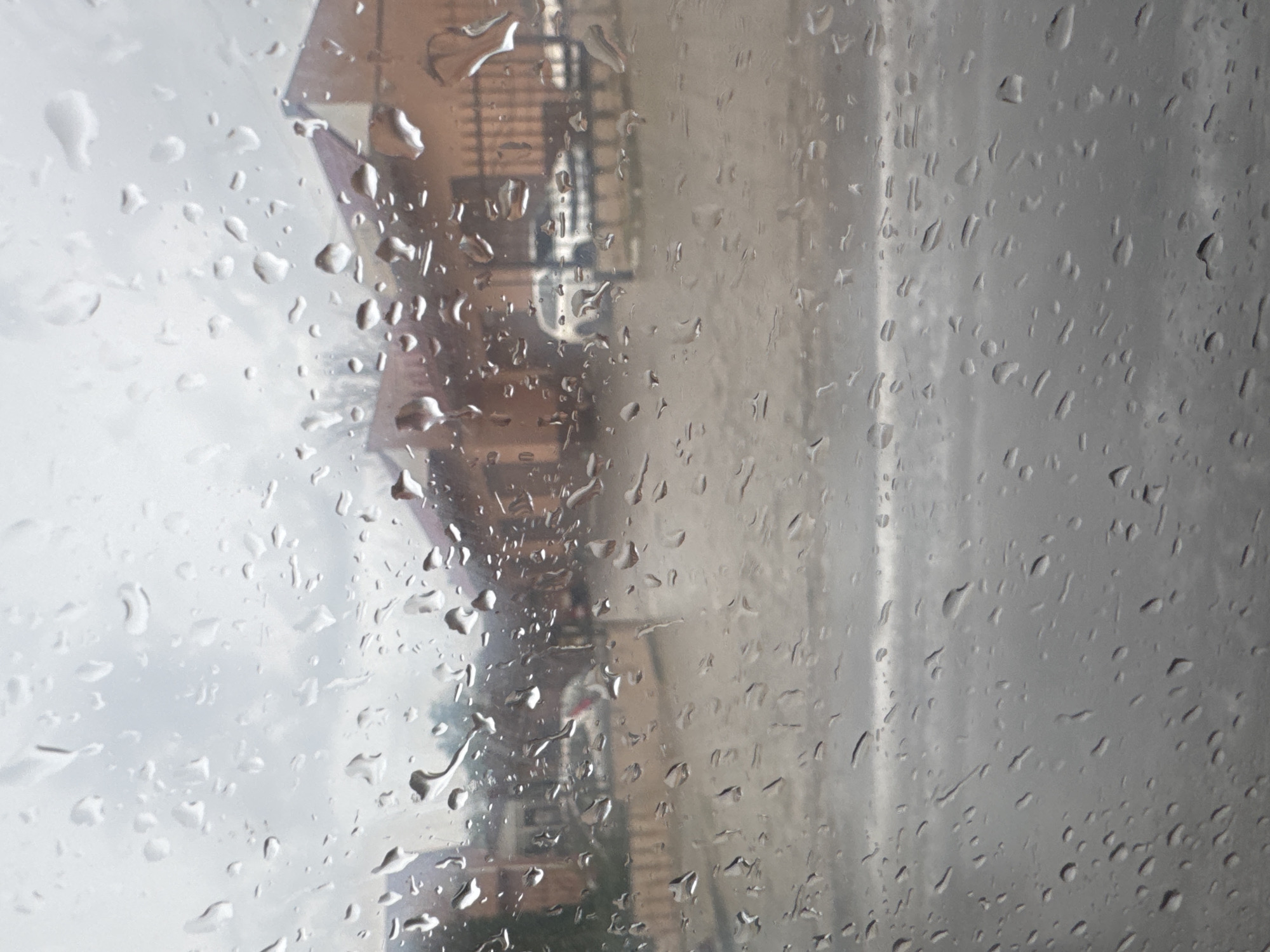 The image shows a view through a window covered in raindrops. In the background, there are buildings with reddish-brown roofs and a black metal fence. A white car is parked in front of one of the buildings. The ground appears wet, suggesting it is raining. The sky is overcast and gray. The scene is likely a residential or commercial area in Tiraspol, Moldova, given the overall aesthetic and architectural style.