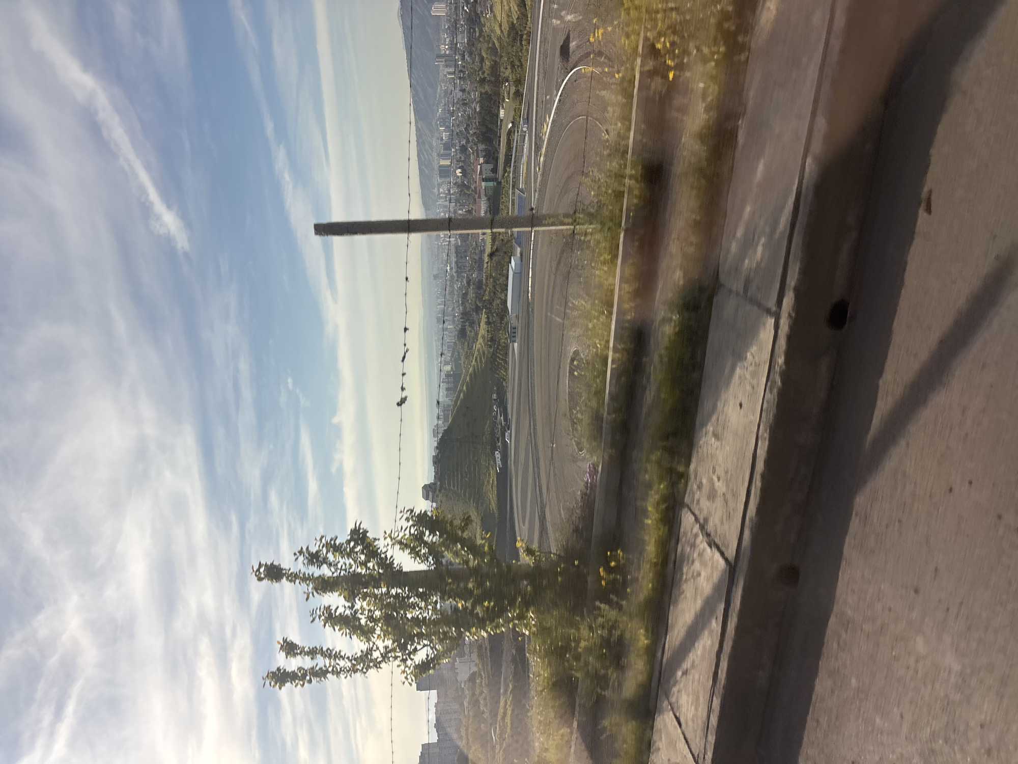 The image, shot from an elevated position possibly inside a vehicle looking out a window, shows a cityscape in Tbilisi, Georgia. In the midground is a multi-lane circuit road with a series of tight curves. Buildings are interspersed throughout the valley below. In the foreground, a tree obscures a portion of the view. A concrete barrier is prominent on the right, casting a shadow on the adjacent walkway. Above, the sky is partly cloudy. A utility pole with a barbed-wire fence is also visible.