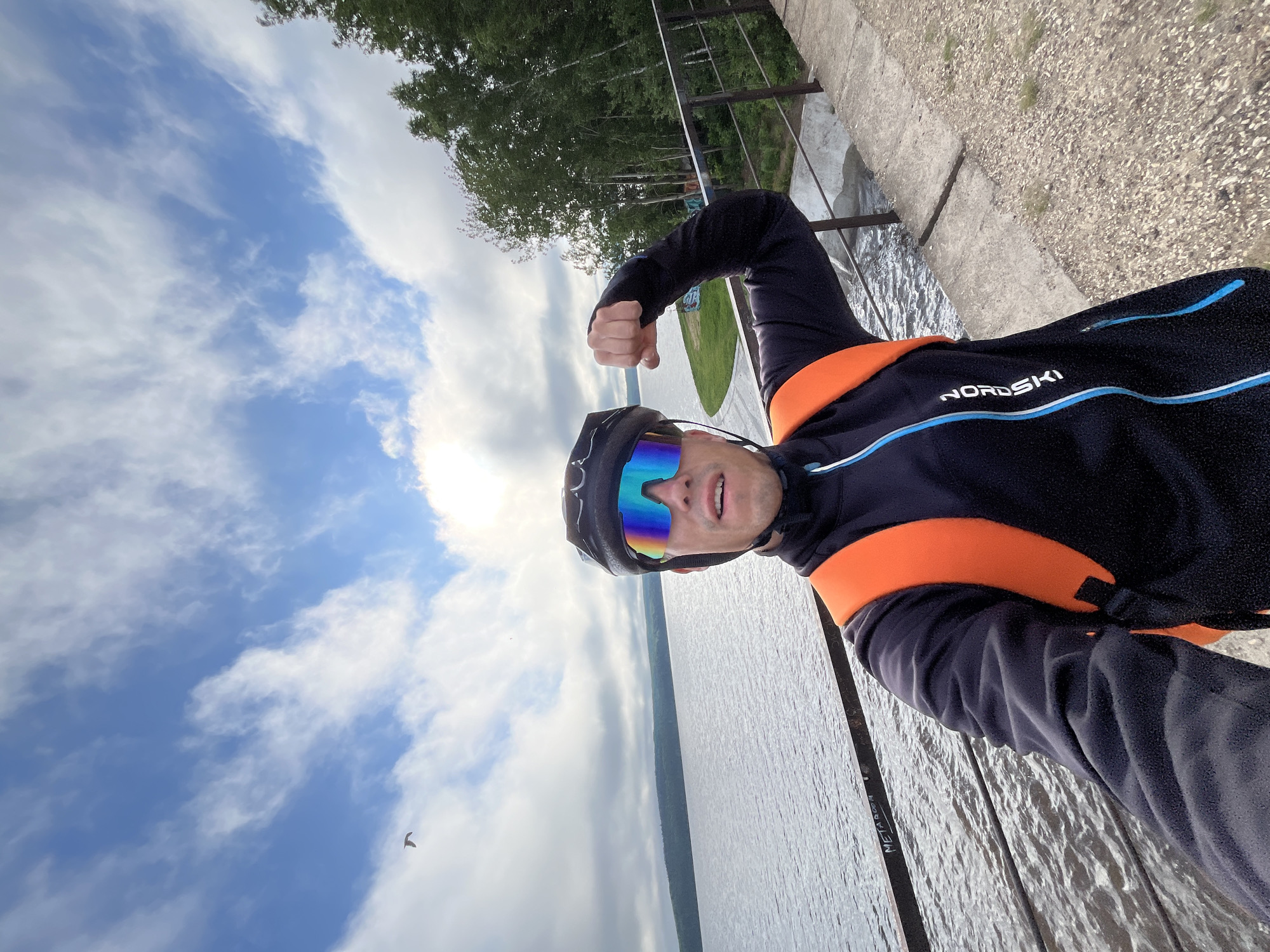 A man in cycling gear, including a helmet and brightly colored goggles, takes a selfie on a concrete walkway next to a body of water. He is wearing a black cycling jacket with an orange backpack. His right arm is flexed, showing his bicep. Behind him, a calm lake stretches to the horizon, with a line of trees on the far bank. The sky is partly cloudy with some blue sky visible. The walkway appears to be part of a dam or bridge structure, with a metal railing and a drop-off to the water below. Graffiti is visible on the railing. The man is likely in Chaikovsky, Russia, as evidenced by the presence of the Kama River reservoir, which is a prominent feature of the city.