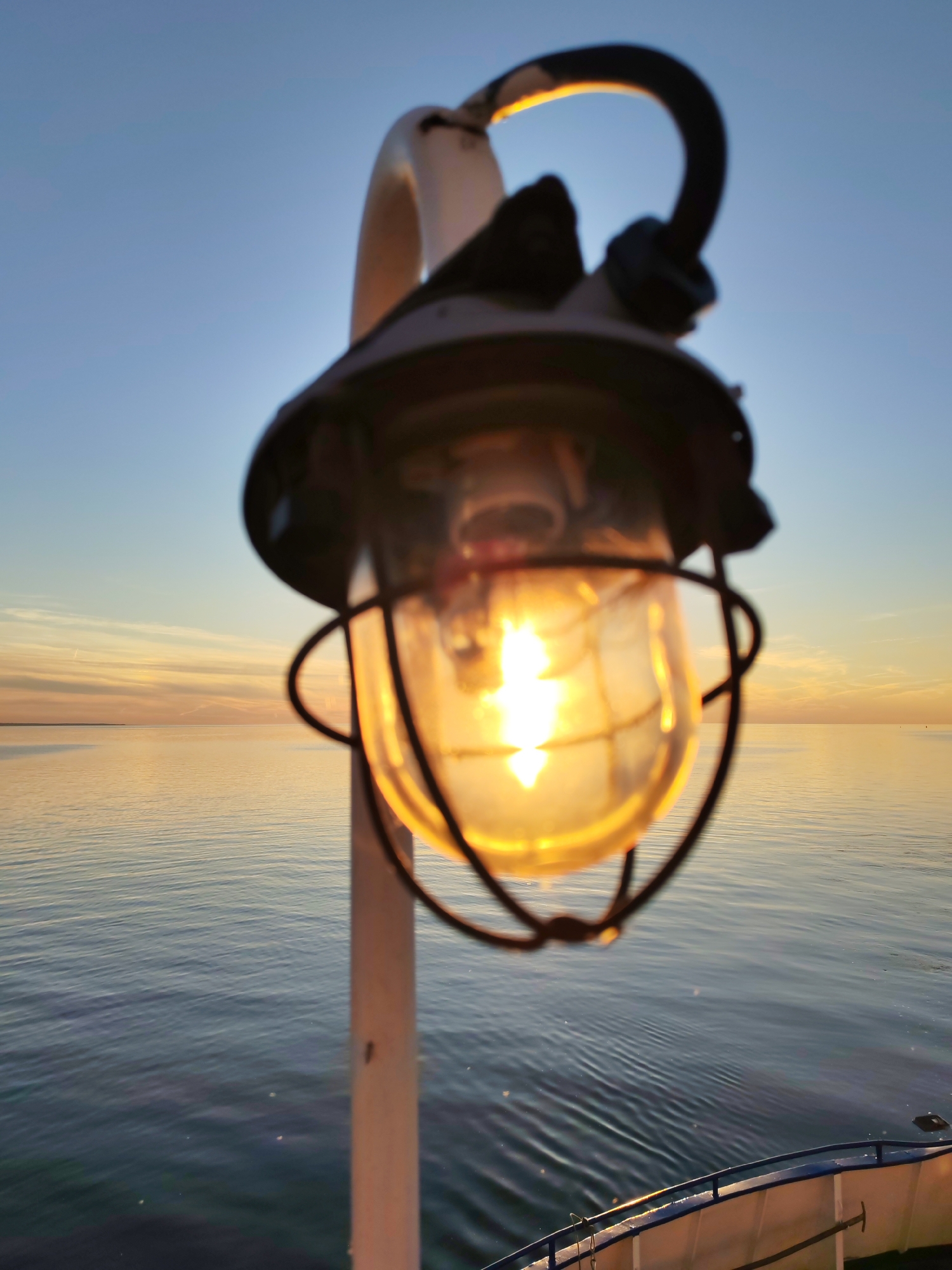 A prominent maritime-style lantern, mounted on a light-colored vertical pole, is centrally positioned in the foreground, illuminated and glowing brightly. The lantern features a dark metal cage enclosing a clear glass globe with a visible incandescent filament. The light from the bulb creates a strong orange-yellow glow, reflecting on the glass. The background is softly blurred but depicts an expansive body of calm water, likely an ocean or large lake, extending to a flat horizon line. The sky above transitions from a pale blue at the top to soft golden and orange hues near the horizon, indicative of either sunrise or sunset. Wispy clouds are present in the sky. Gentle ripples are visible on the water's surface, reflecting the sky's colors. In the lower right corner, a segment of a boat's white hull and blue railing is visible, indicating the perspective is from on board a vessel. The location is on open water, in the city of Unknown, Unknown.